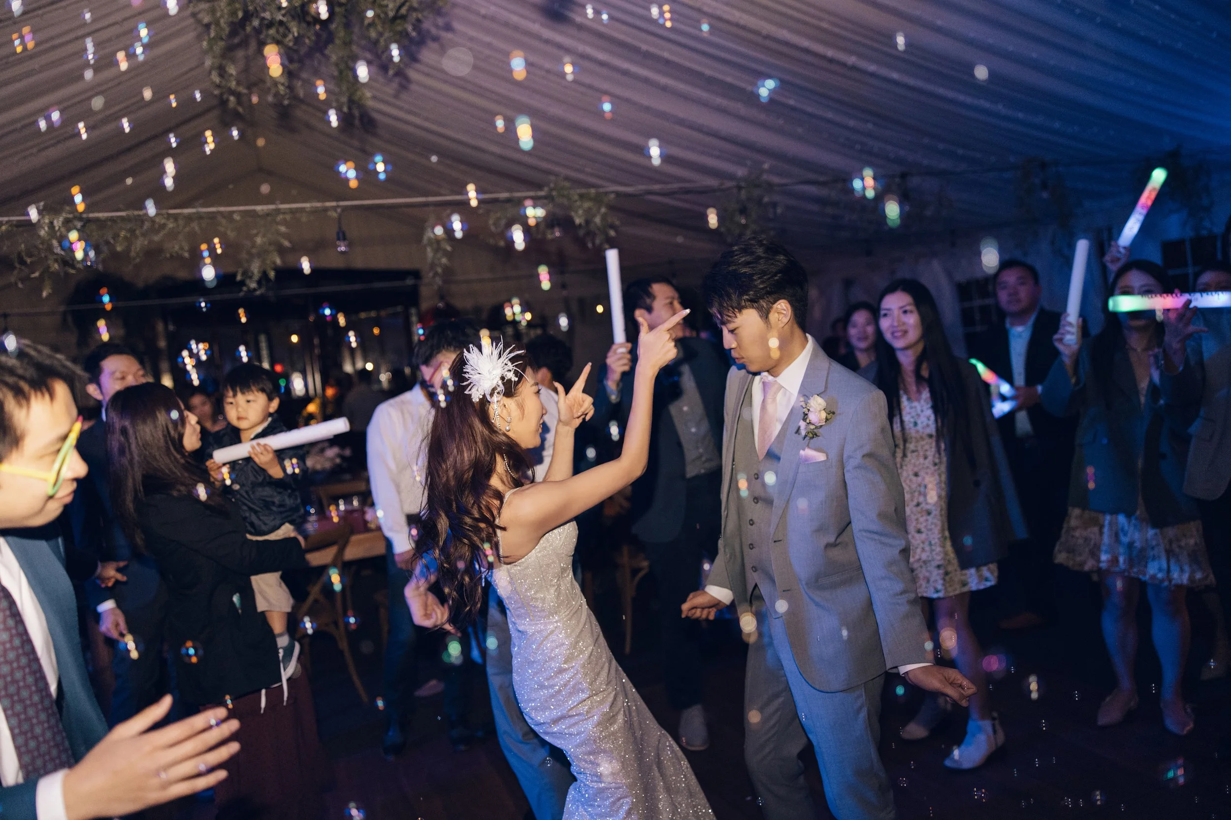 A bride and groom dancing at their wedding reception with guests around them, some holding glowing sticks, under a decorated ceiling with hanging lights.
