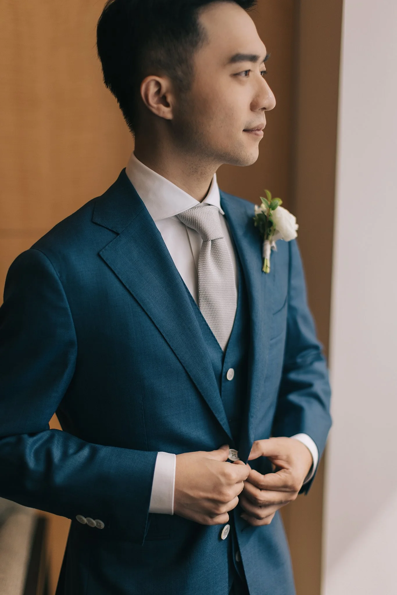 A groom in a blue suit with a white shirt and silver tie, adjusting his jacket, standing near a window.