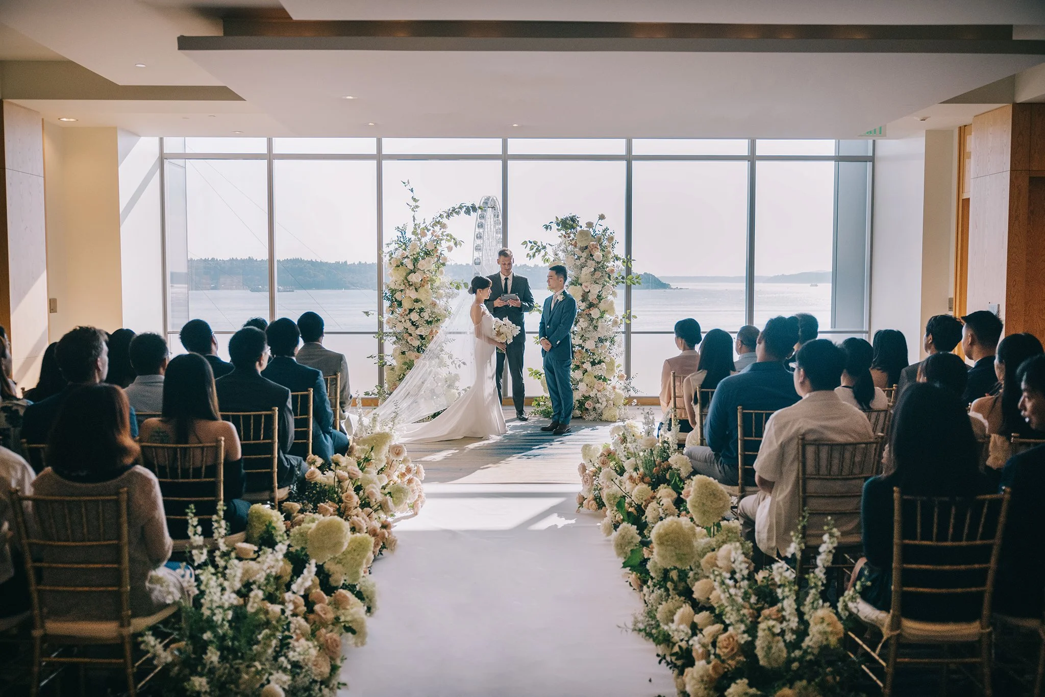 A wedding ceremony taking place indoors by large windows overlooking water and a Ferris wheel, with a bride and groom standing before an officiant, surrounded by white floral arrangements.