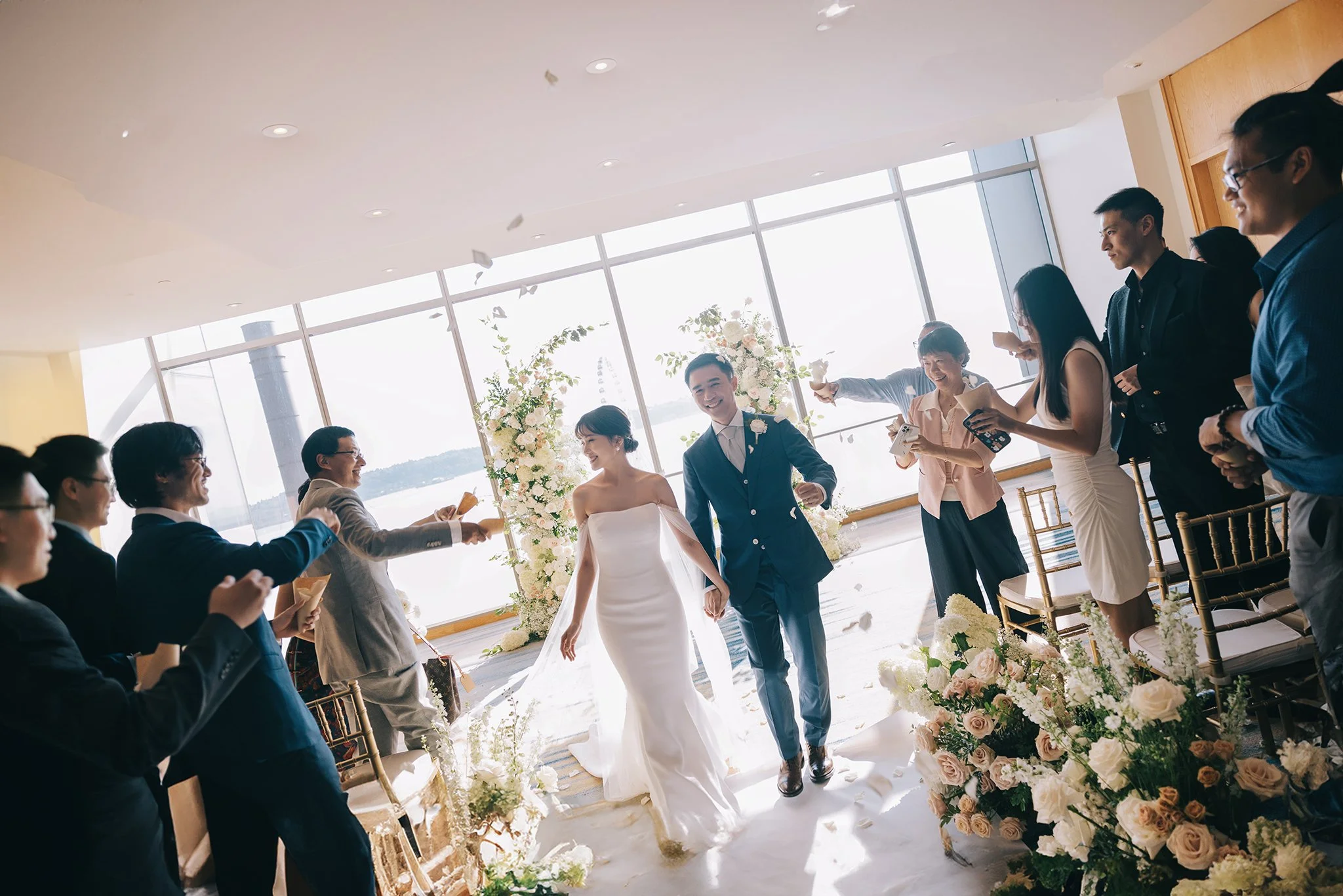 Bride and groom walking down aisle in a bright, decorated wedding venue, surrounded by friends and family celebrating with flower petals and smiling.