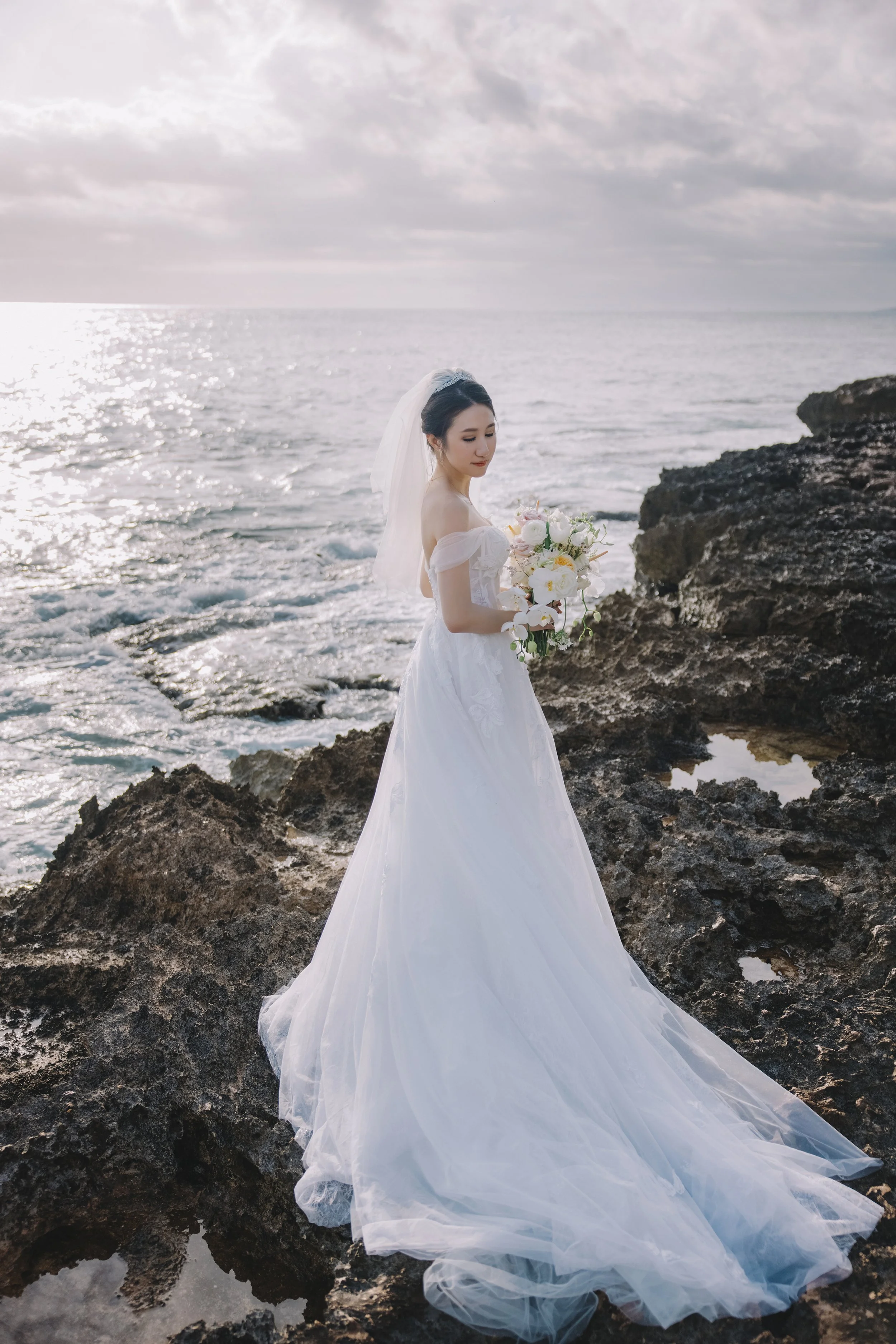 Bride in a white wedding gown holding a bouquet of flowers stands on rocks by the ocean at sunset.