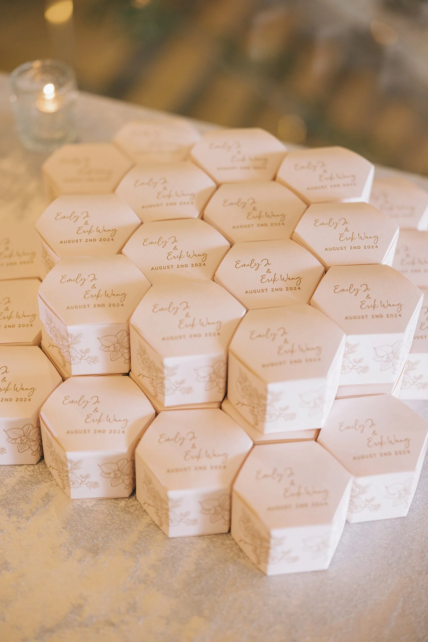Hexagonal white favor boxes with floral designs and personalized text, stacked on a table, with a small candle holder in the background.