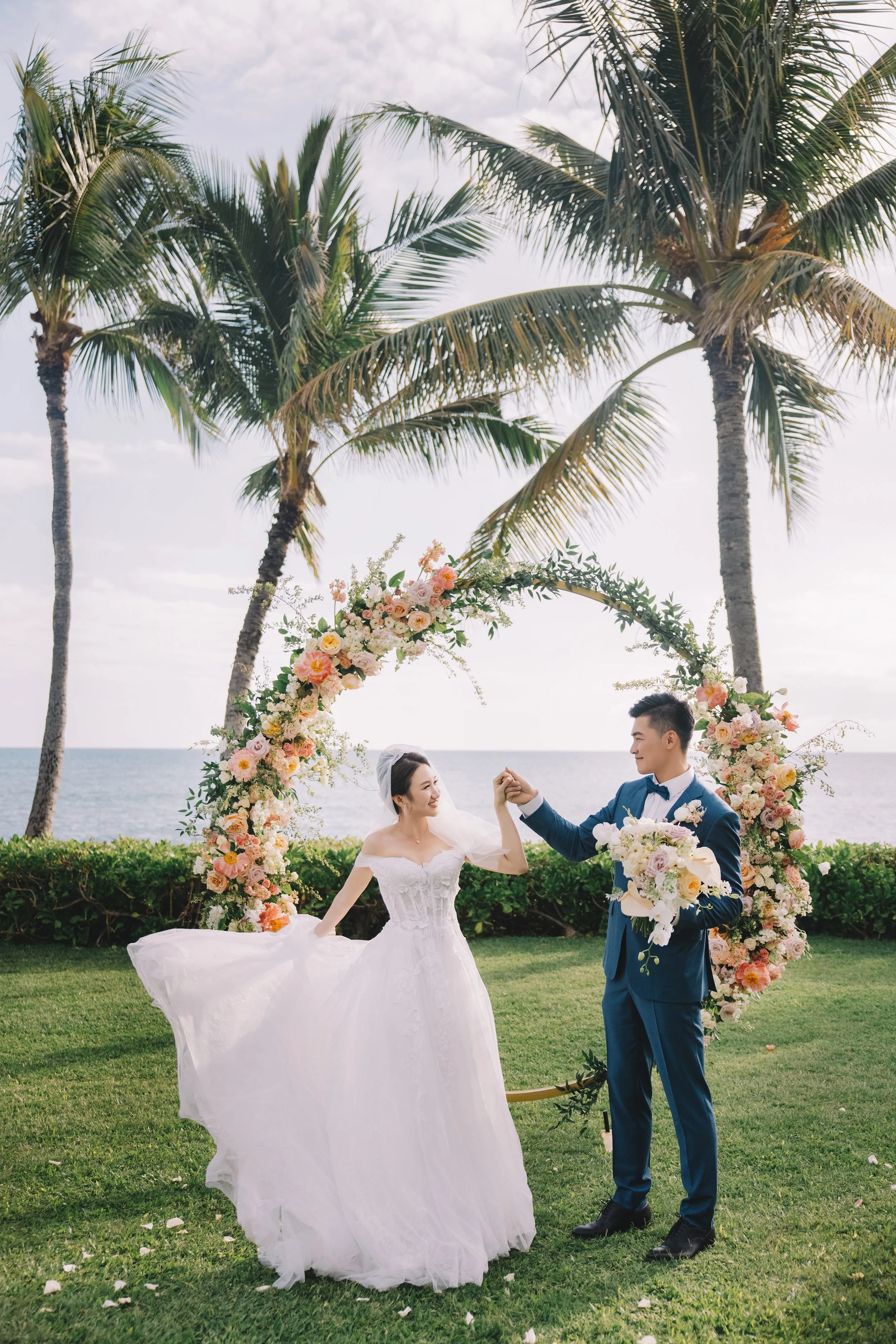 A bride and groom dancing outdoors on a grassy area with a floral arch, palm trees, and ocean in the background.