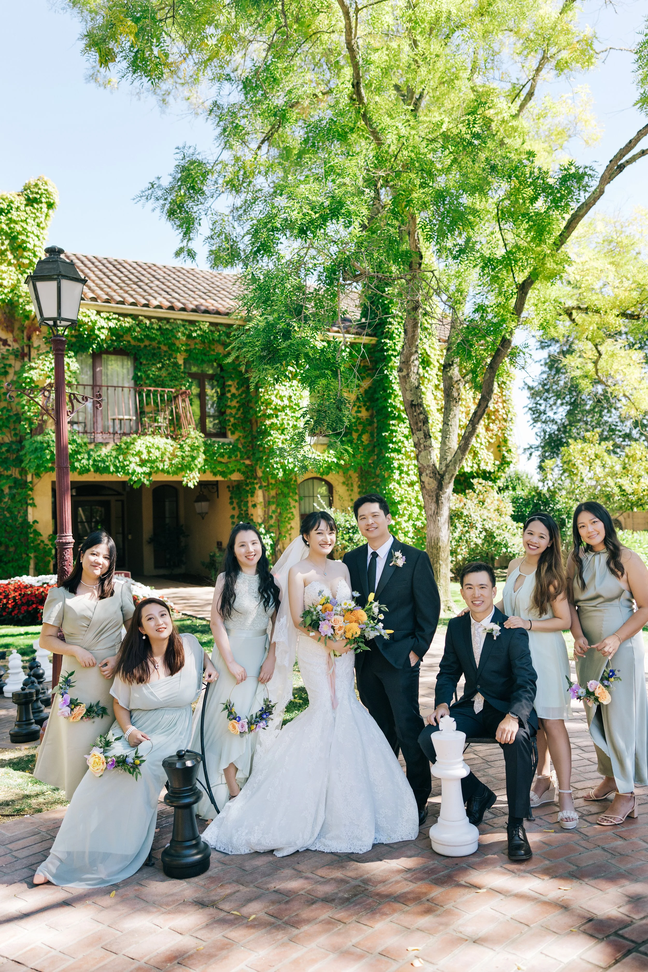 Group of people at a wedding, with a bride in a white gown holding a bouquet and surrounded by friends in light-colored dresses and suits, in an outdoor garden setting with large trees and a house covered in greenery.