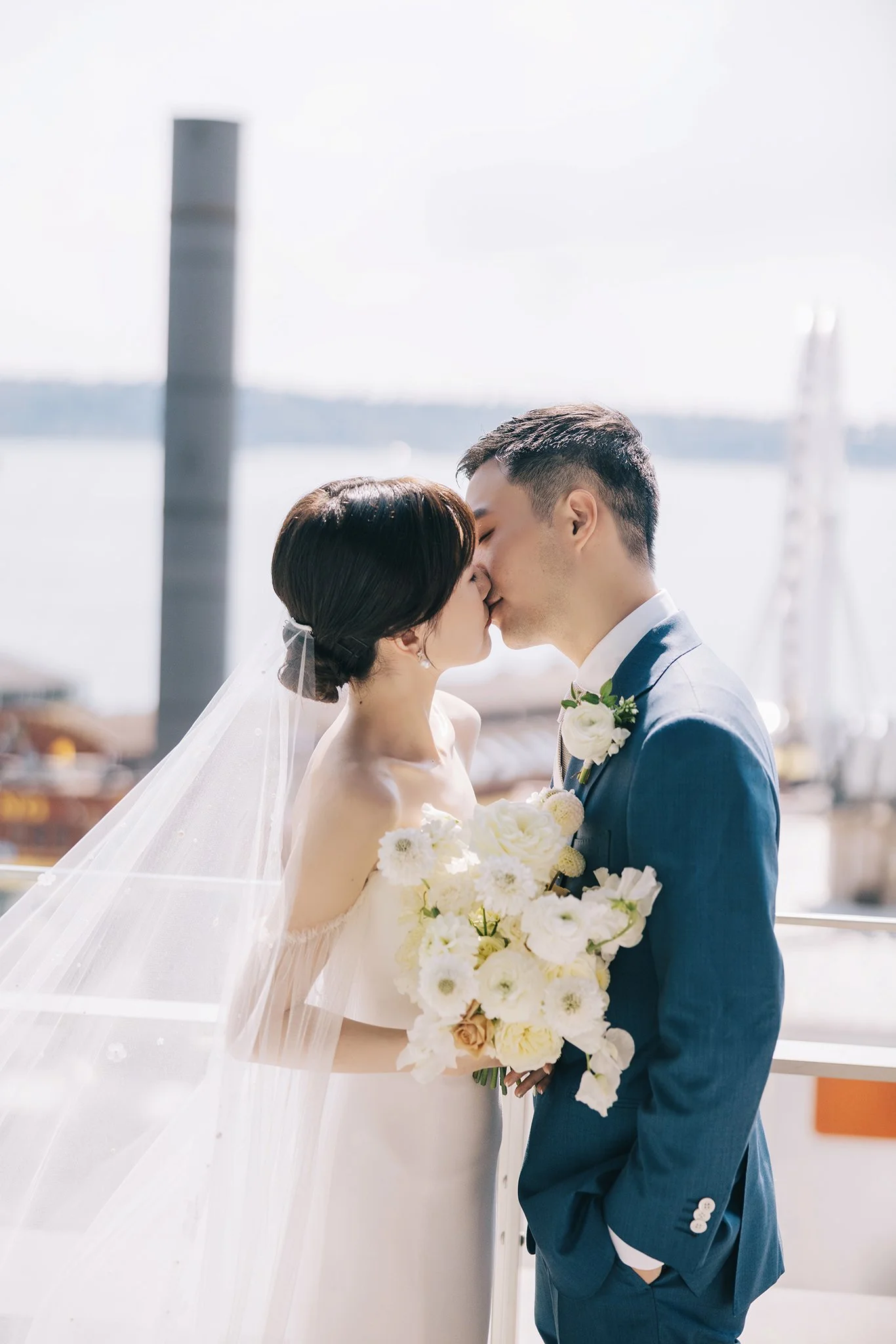 A bride and groom sharing a kiss during their wedding, with a waterfront and cloudy sky in the background, the bride in a white dress holding a bouquet of white flowers and the groom in a blue suit with a white boutonniere.