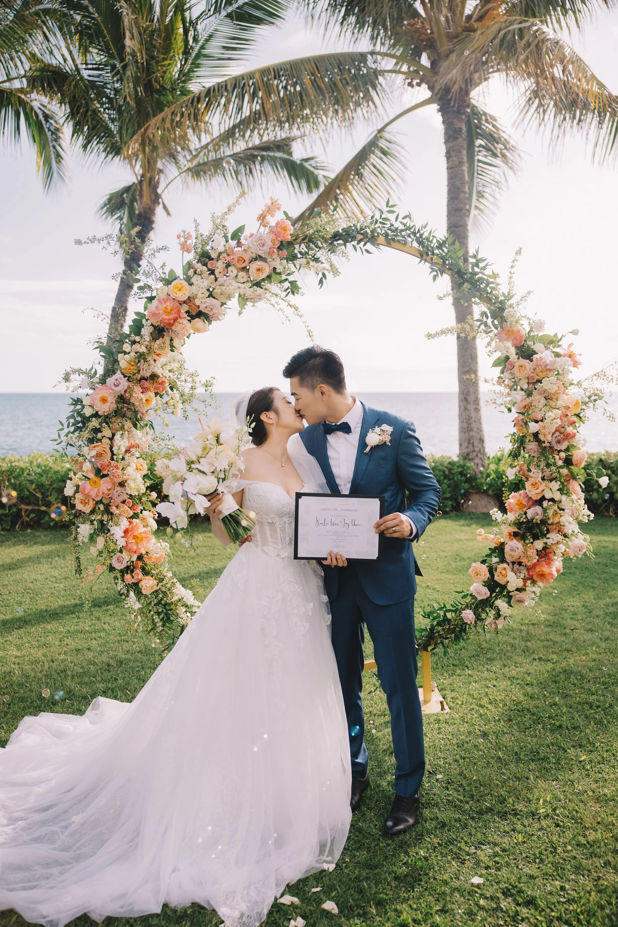 A newlywed couple shares a kiss during their wedding ceremony on a grassy area near the ocean. They are positioned under a floral arch with pink and white flowers, and there are tall palm trees in the background.