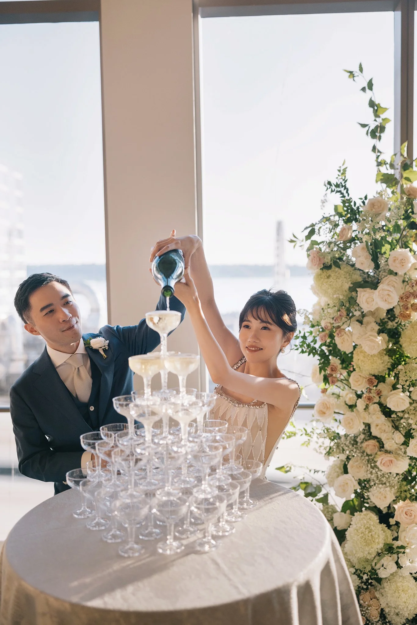 A bride and groom pouring champagne into a pyramid of glasses during their wedding celebration, with a large floral arrangement nearby and a window showing a river view in the background.