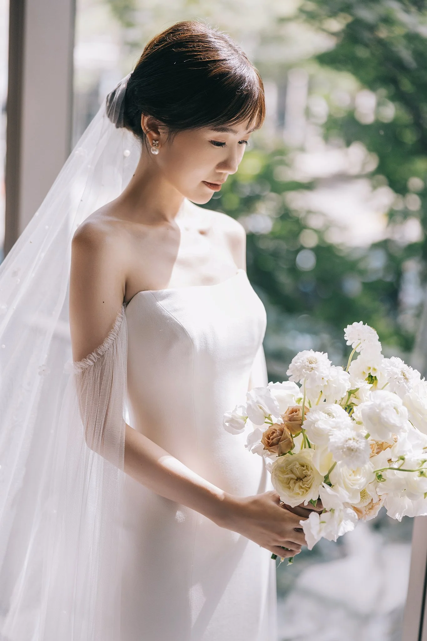 A bride in a white wedding dress holding a bouquet of white and brown flowers, standing near a window with trees outside.