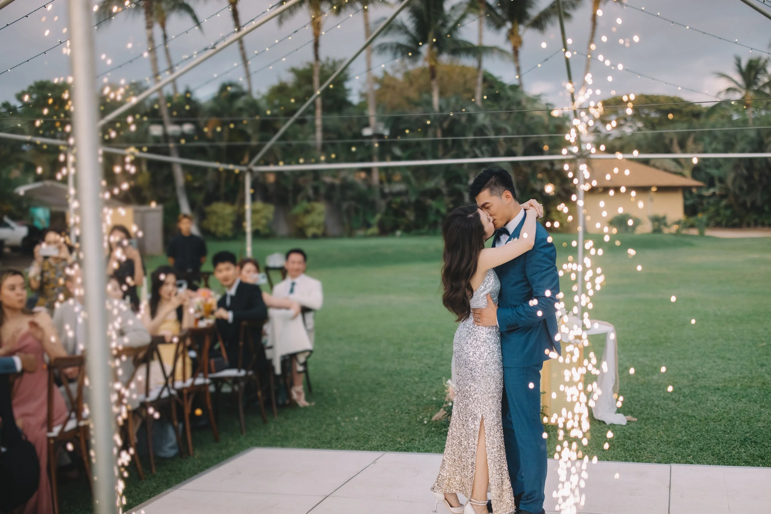 A couple is kissing during their wedding reception under string lights and sparklers, with seated guests and a lush outdoor setting in the background.