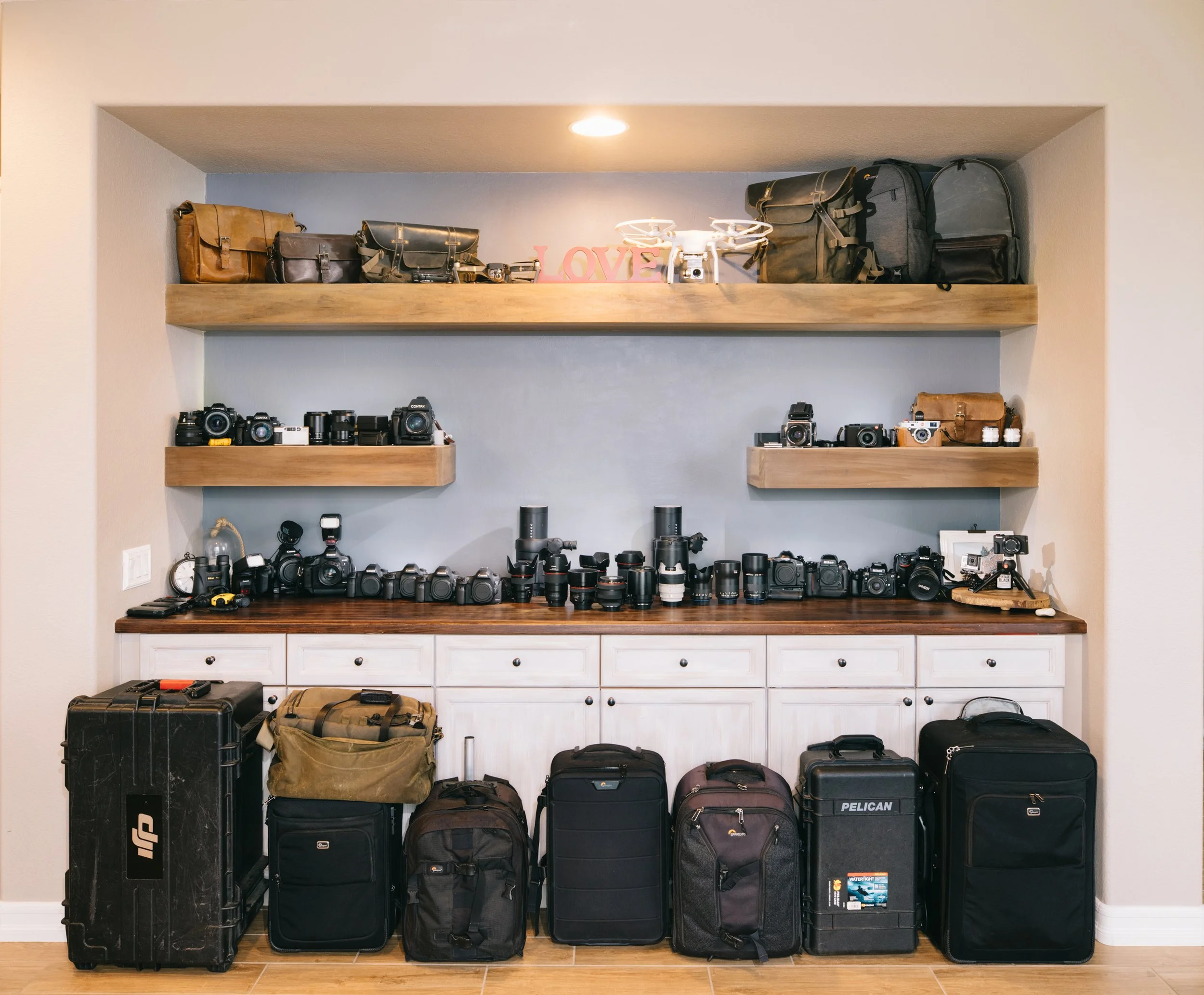 A collection of cameras, camera lenses, and photography equipment organized on shelves and a countertop, with travel cases and backpacks lined up below.