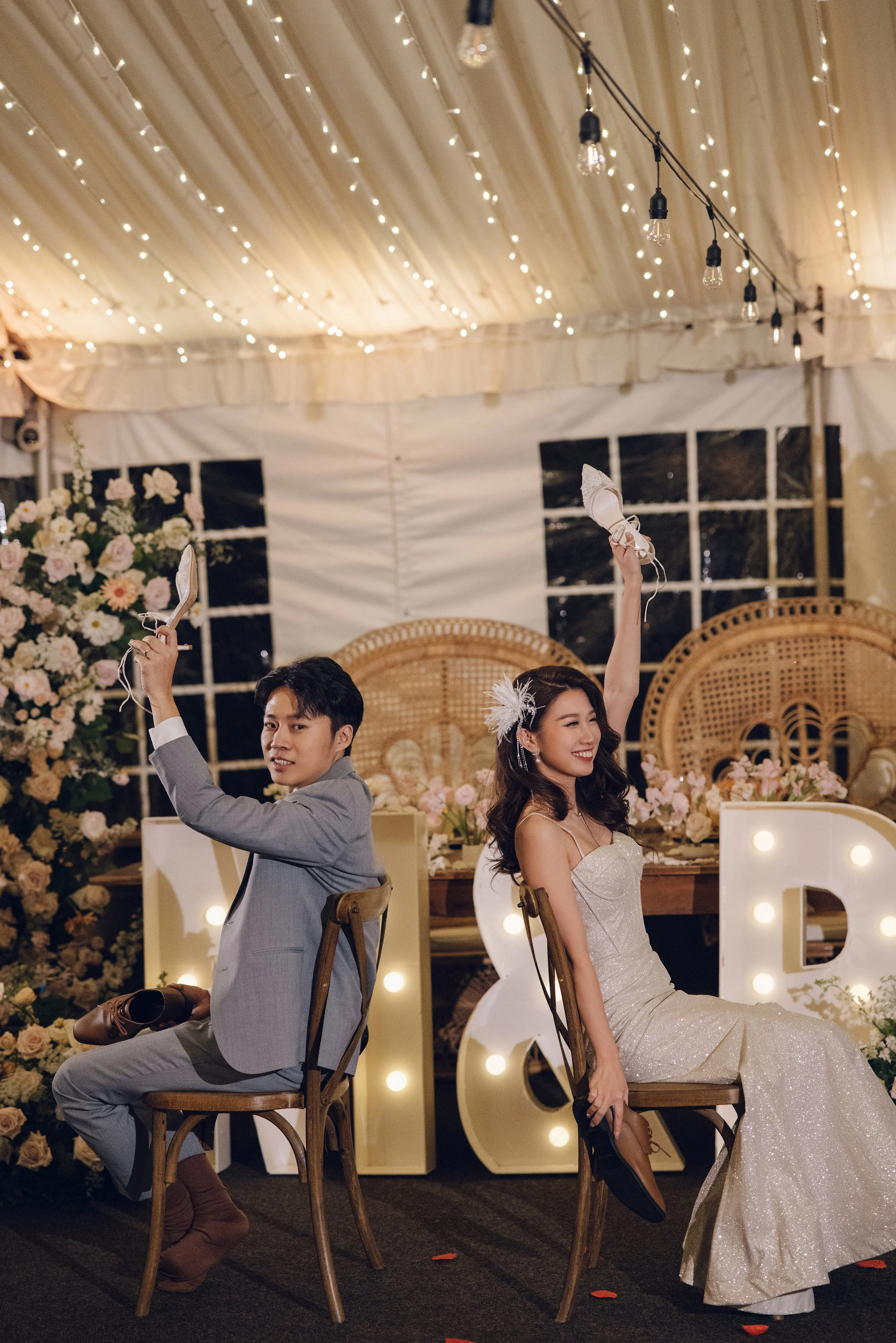 A bride and groom sit on chairs back to back inside a decorated wedding tent, each holding one of a pair of bridal shoes raised in the air, smiling and enjoying the moment during their wedding celebration.