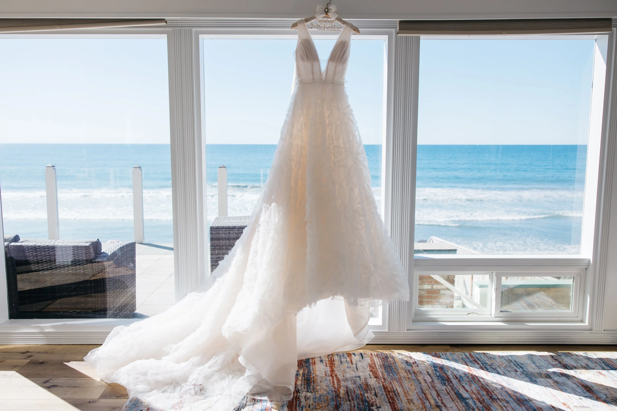 A white wedding dress hanging in front of large windows with a view of the ocean and a balcony.