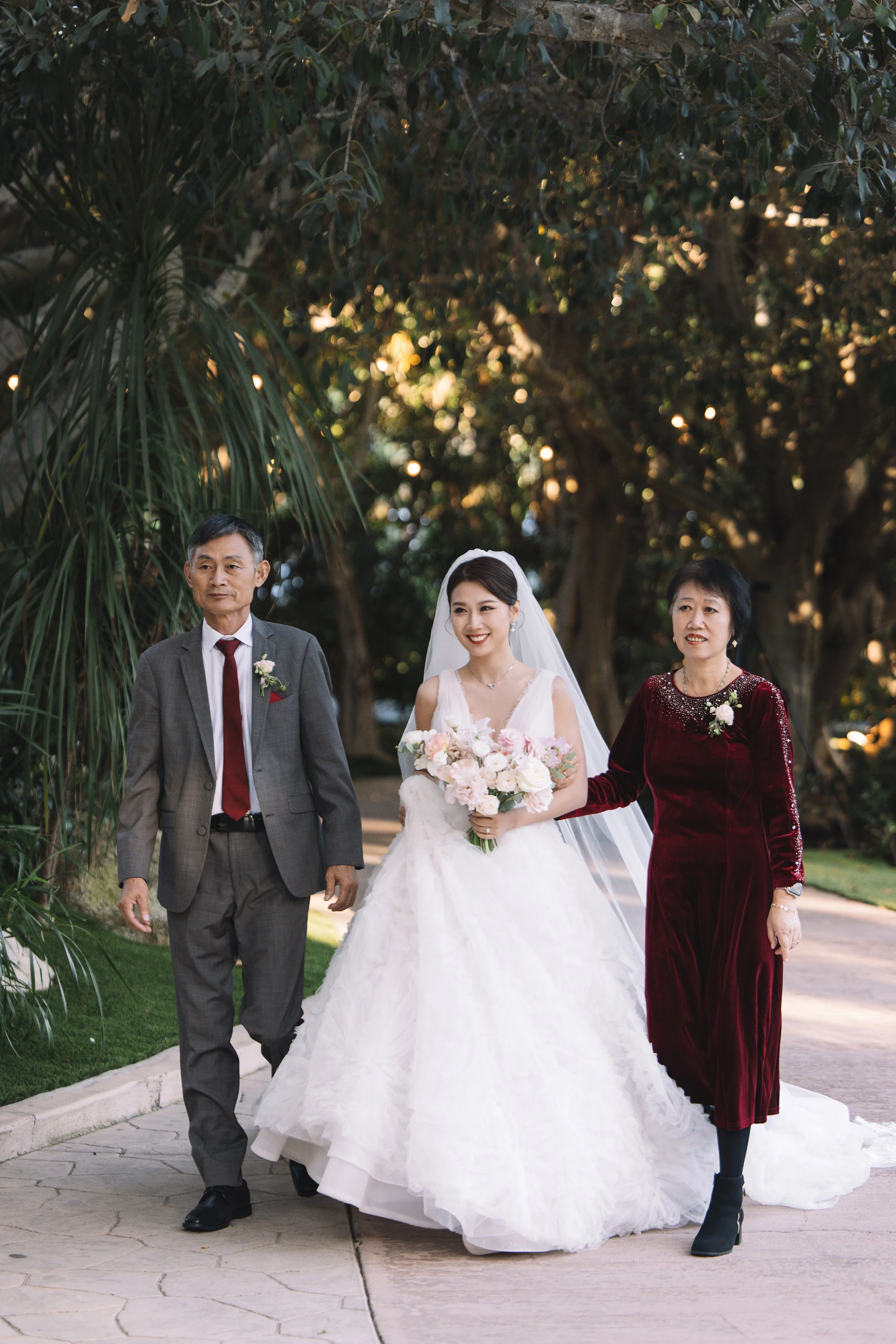 A bride in a white wedding gown holding a bouquet of flowers walks with her parents outdoors on a paved pathway, surrounded by trees. The father wears a gray suit with a red tie, and the mother wears a long, velvet maroon dress.