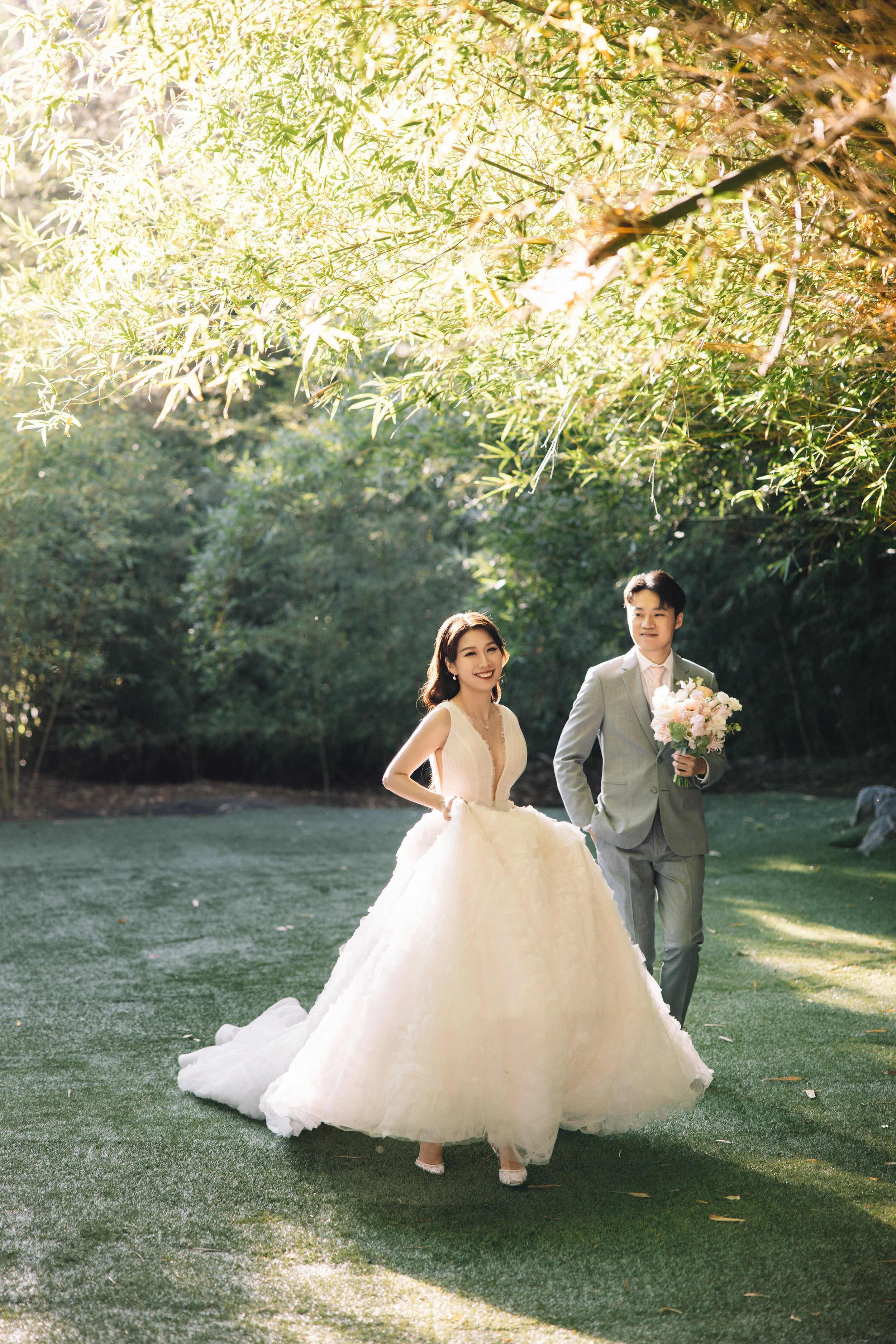 A bride in a white wedding gown and a groom in a light gray suit walking outdoors on a grass path, holding a bouquet of flowers, surrounded by green trees and sunlight.