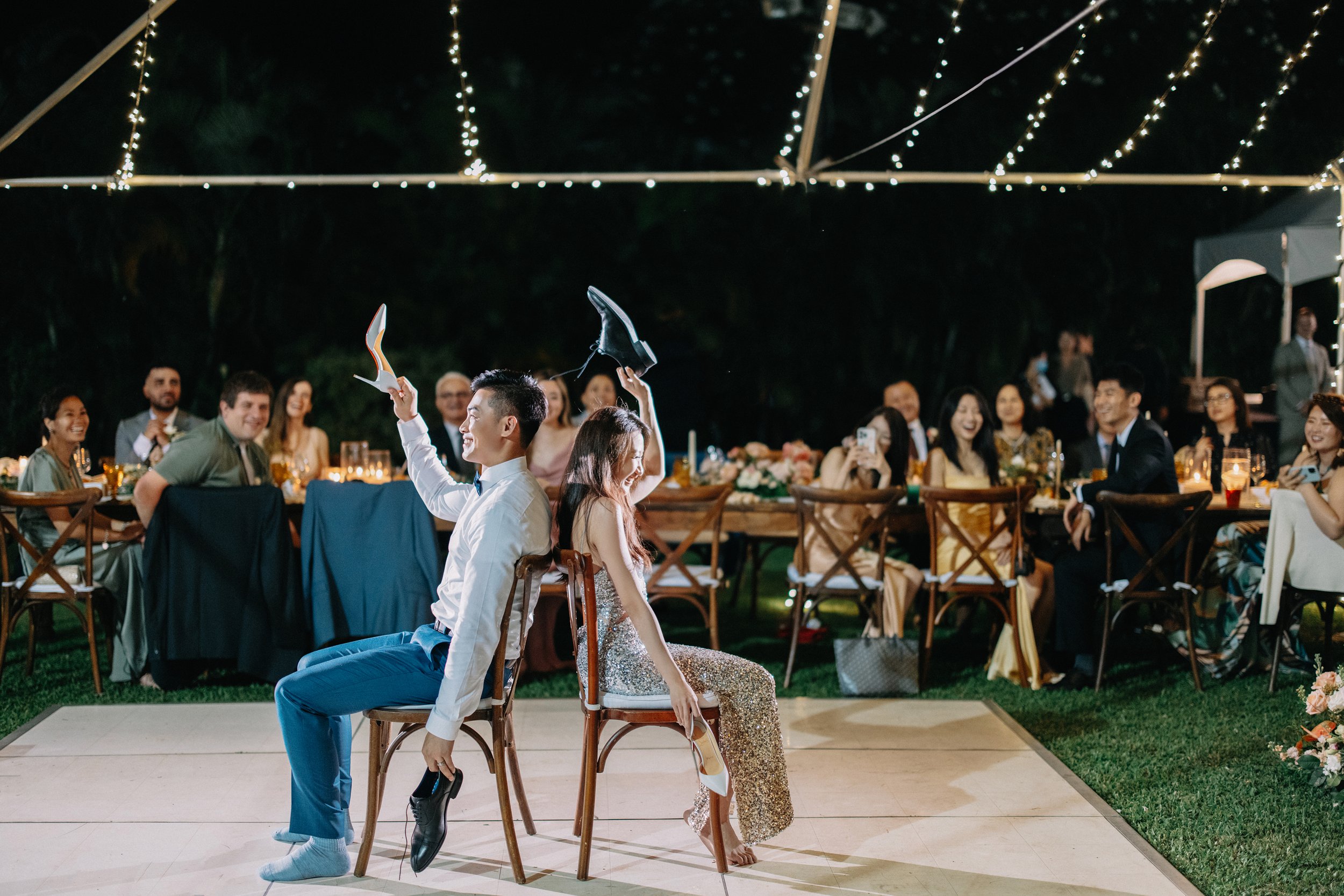 Two people, a man and a woman, seated back-to-back on chairs during a wedding reception, holding shoes above their heads and playing a game. Guests watch and laugh at the event under string lights at night.