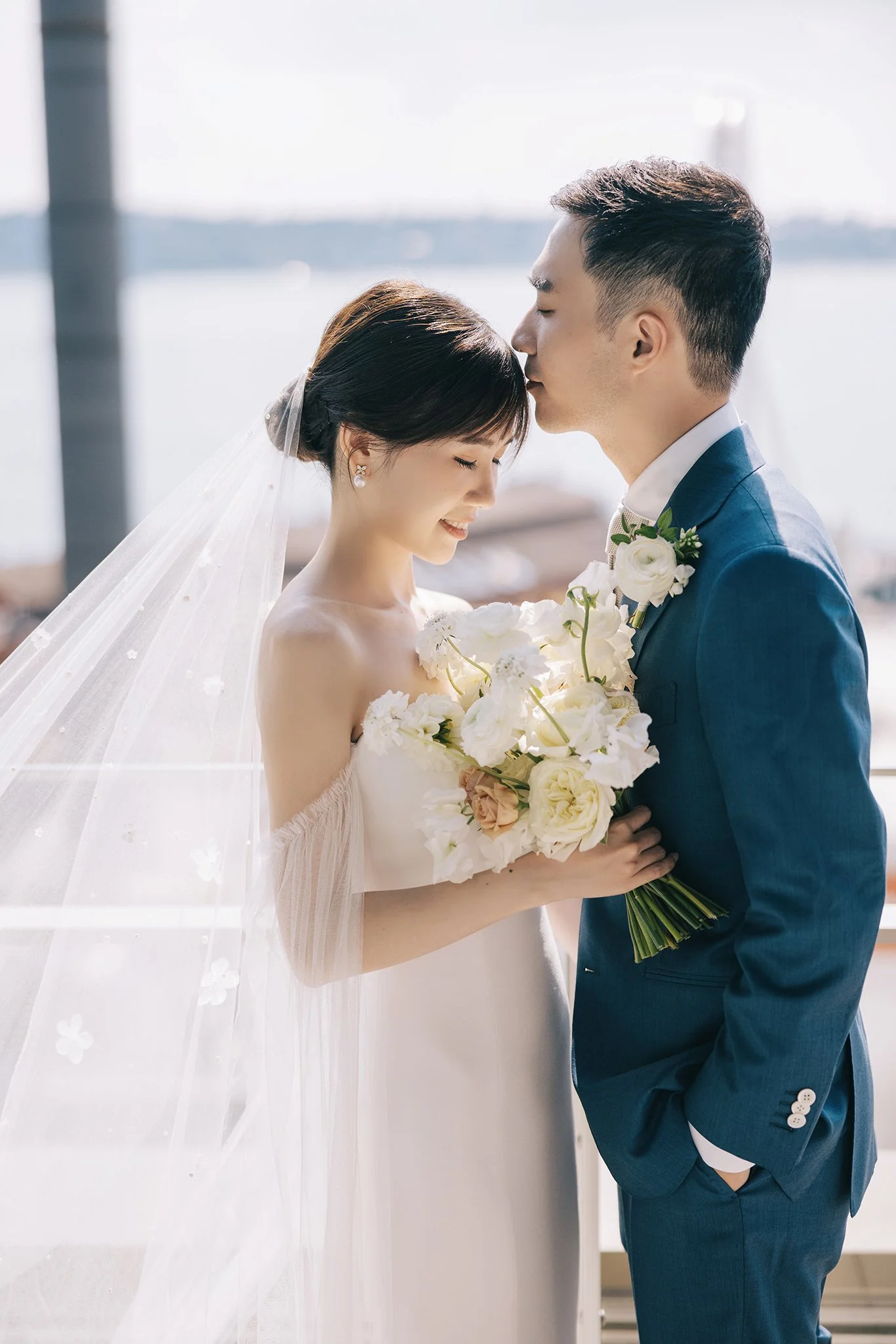 A bride and groom share a tender moment, with the groom kissing the bride's forehead as she smiles with her eyes closed, holding a bouquet of white and blush flowers, on a waterfront background.