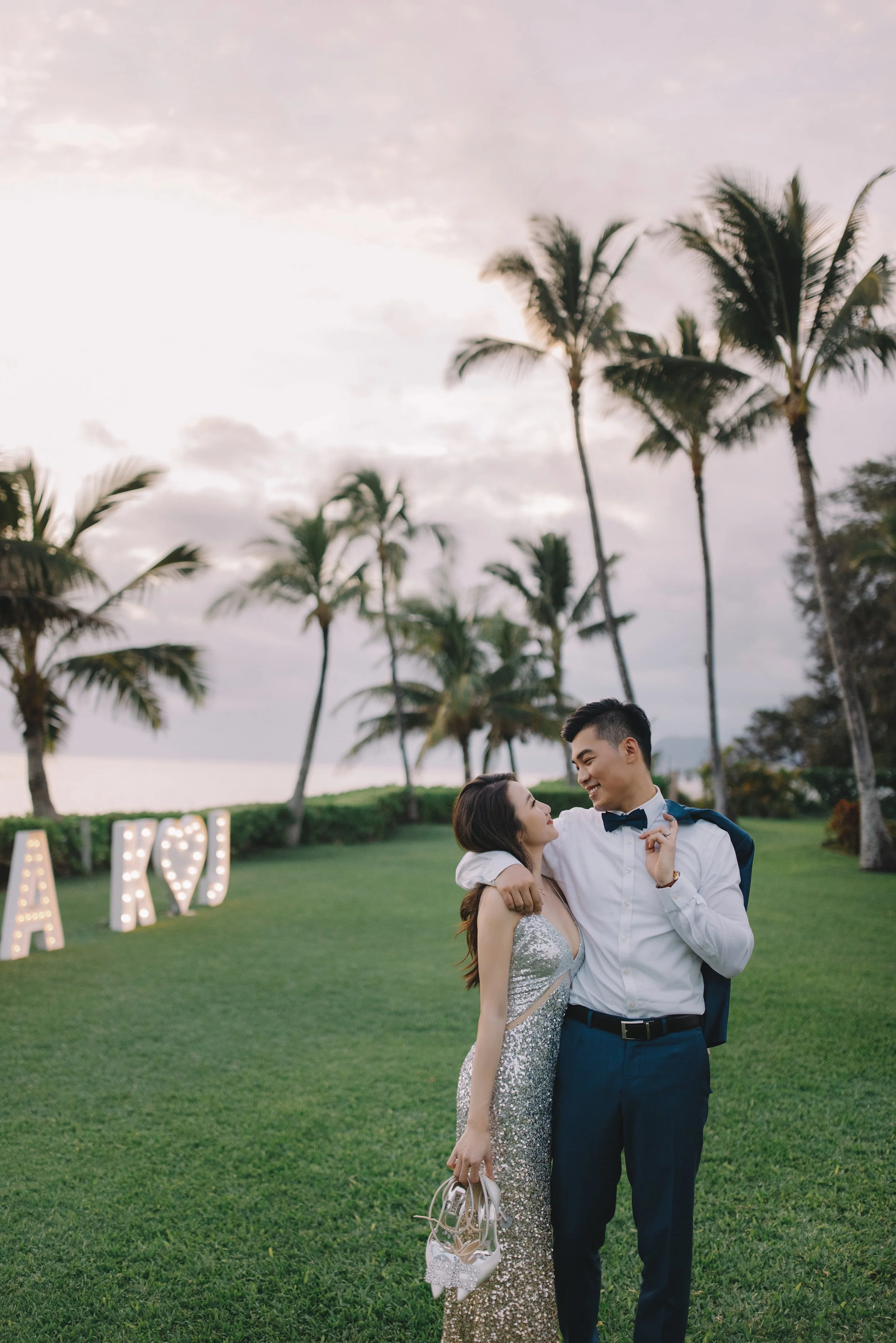 A couple dressed in formal attire enjoying a romantic moment outdoors, with palm trees and lit marquee letters spelling 'LOVE' in the background.