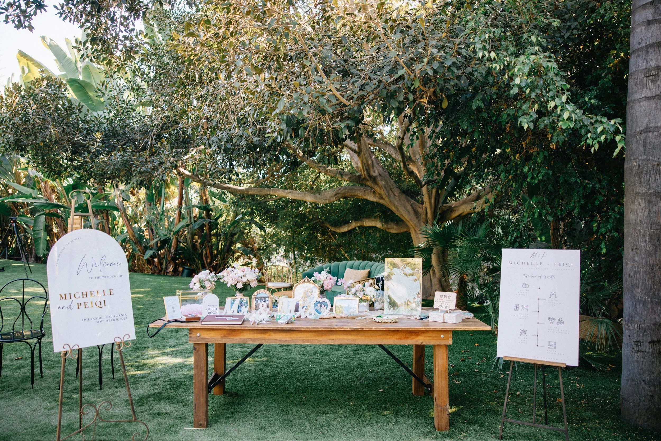 Outdoor wedding reception setup with a wooden table decorated with flowers, picture frames, and signs, surrounded by trees and greenery.