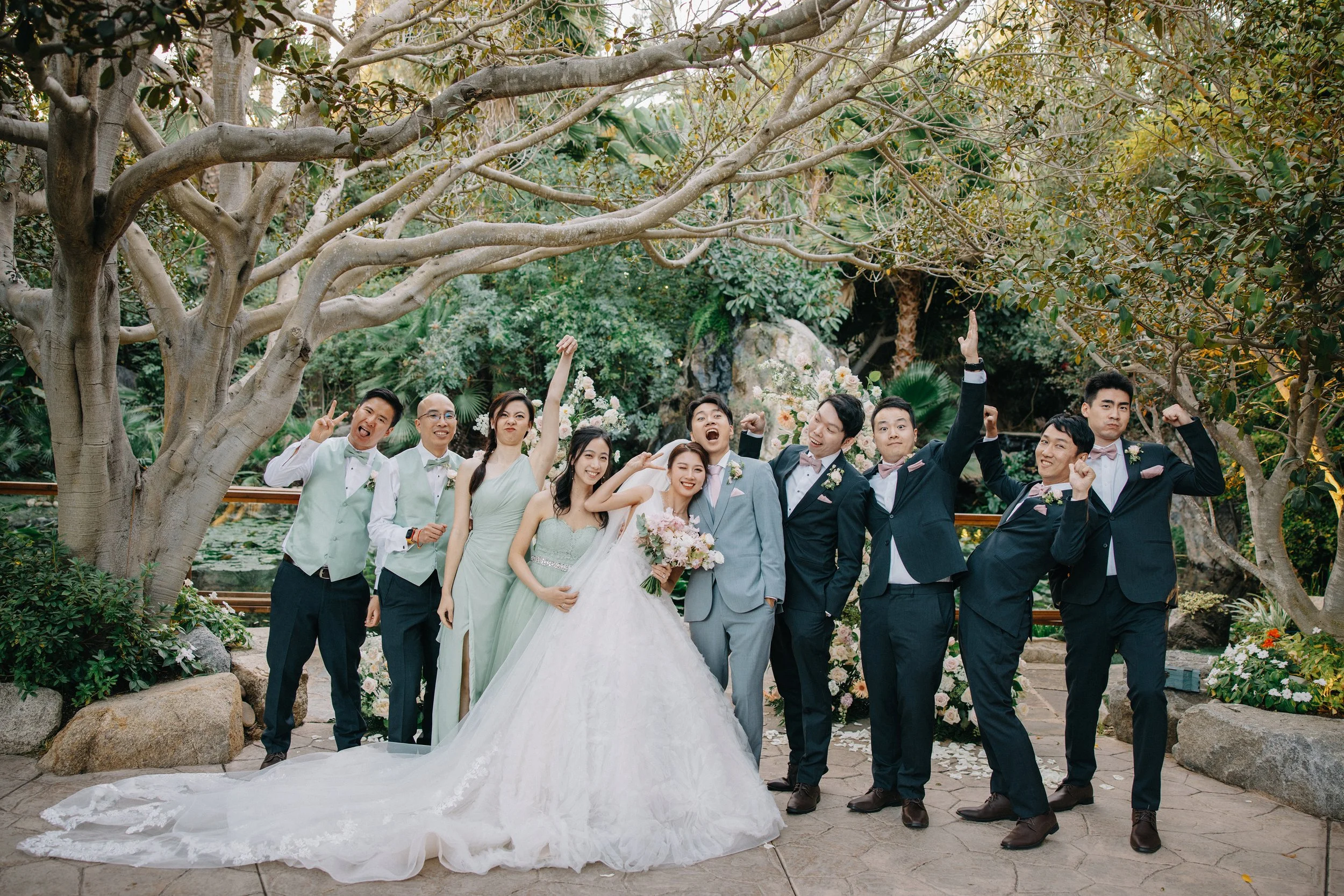 A group of people celebrating a wedding outdoors, with a bride and groom in the center, surrounded by friends in formal attire, with trees and flowers in the background.