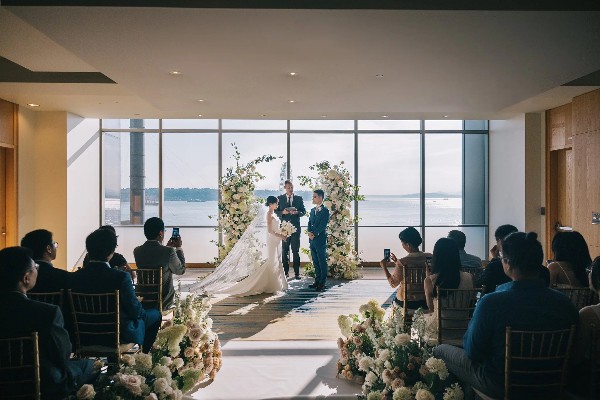 A wedding ceremony taking place in a room with large windows overlooking water, decorated with floral arrangements, with guests seated and taking pictures of the bride and groom at the altar.
