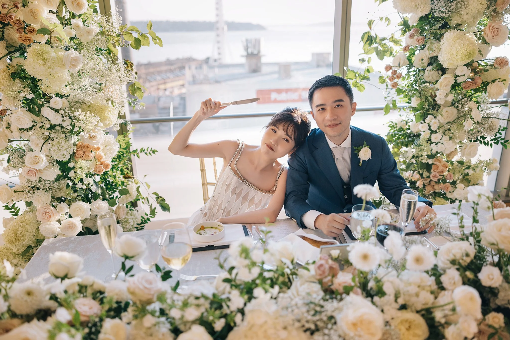 A newlywed couple at their wedding reception, sitting at a decorated table surrounded by white and light pink flowers, with a city waterfront view in the background.