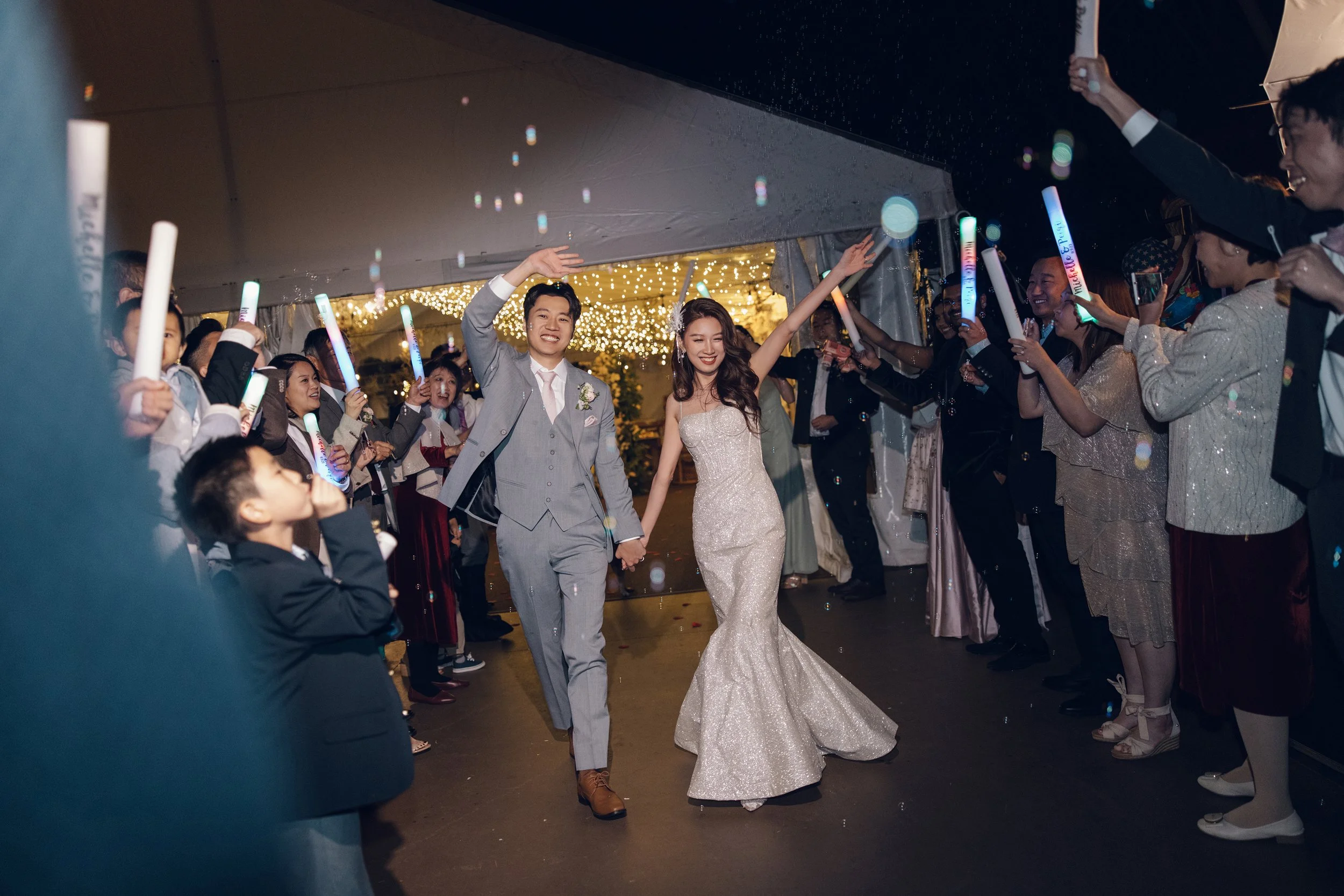 A bride and groom walking hand in hand through a crowd of wedding guests who are celebrating with glow sticks, under a decorated tent with string lights at night.