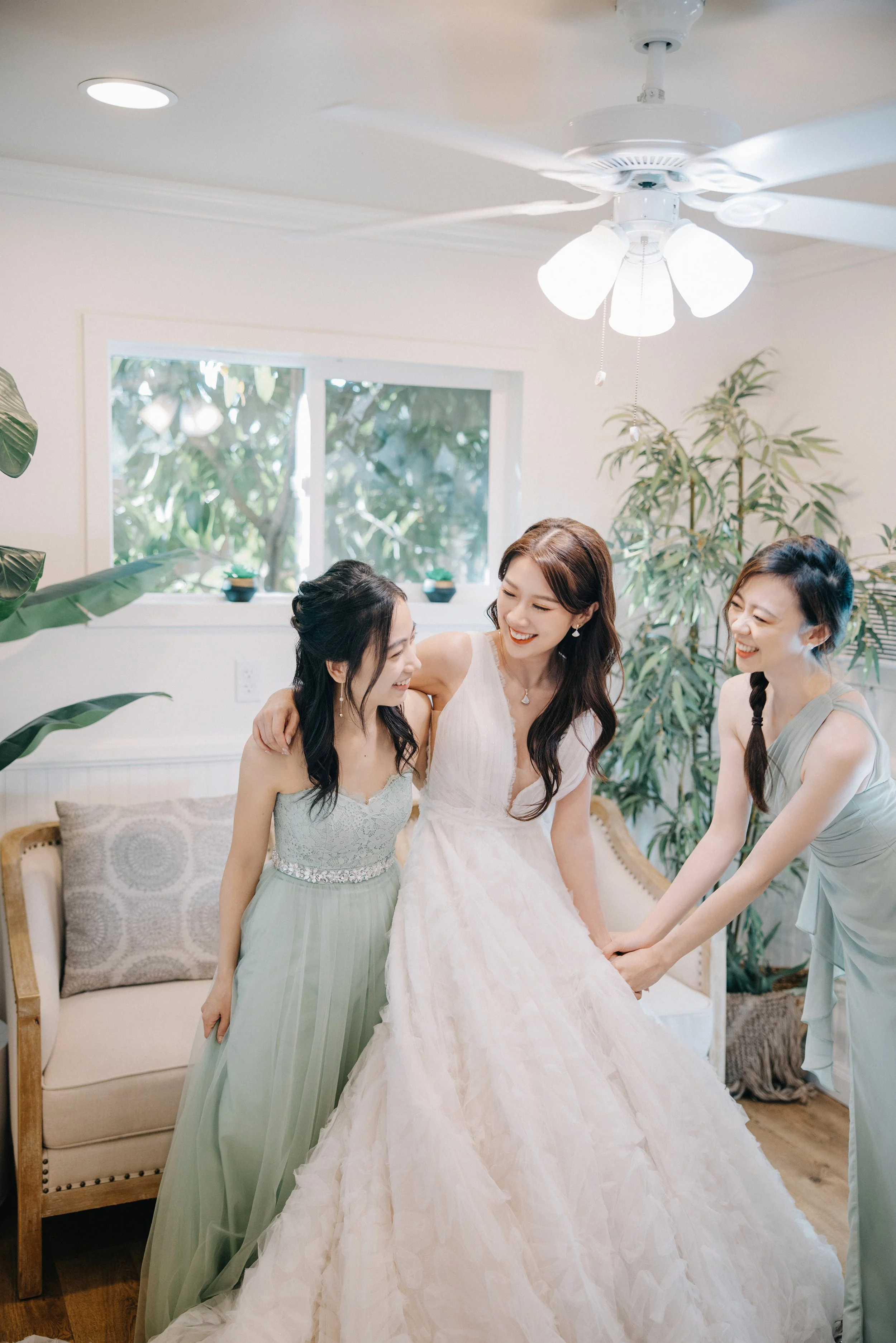 Three women smiling and holding a bride's dress in a bright, green-hued room with plants and a window.