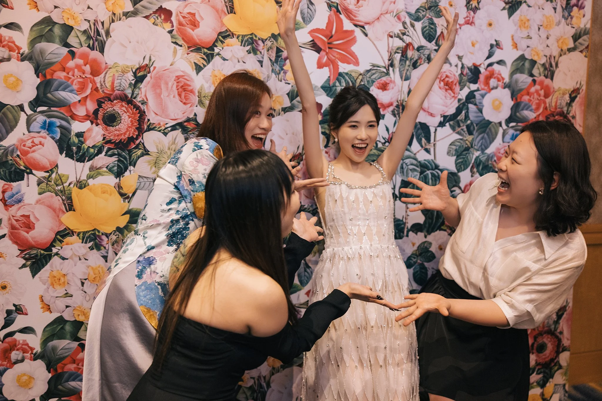 Four women celebrating and laughing in front of a floral background. One woman in a white dress has her arms raised, and the others are reaching out and smiling.
