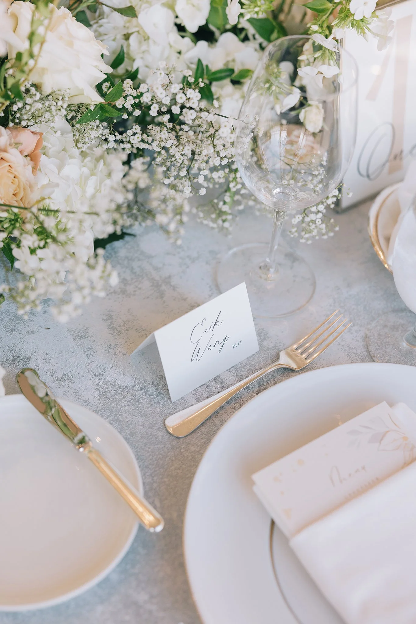 Elegant wedding table setting with white and blush flowers, a wine glass, a gold fork, a white plate with a white napkin, and a place card labeled 'Eric Wang'.