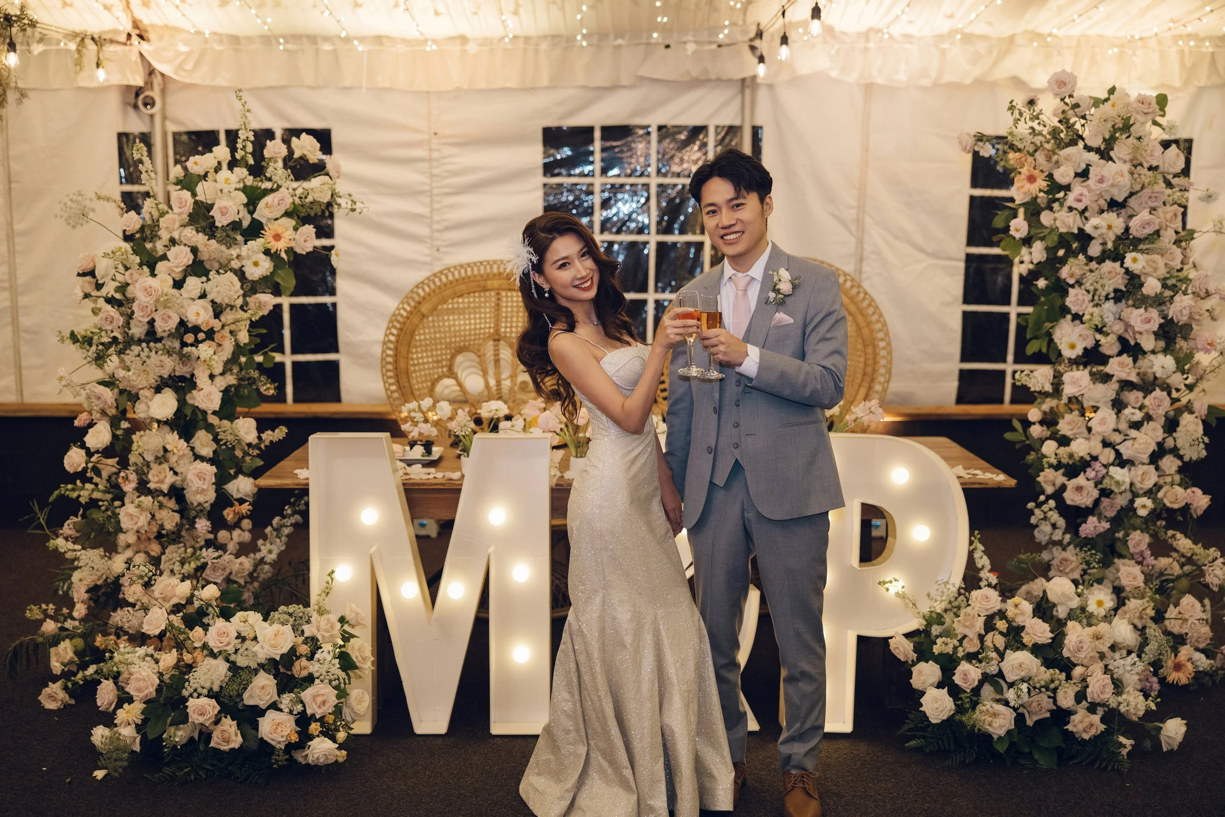 A bride and groom celebrating with champagne in front of large illuminated letters spelling 'M' and 'P', surrounded by floral arrangements in a decorated wedding tent.