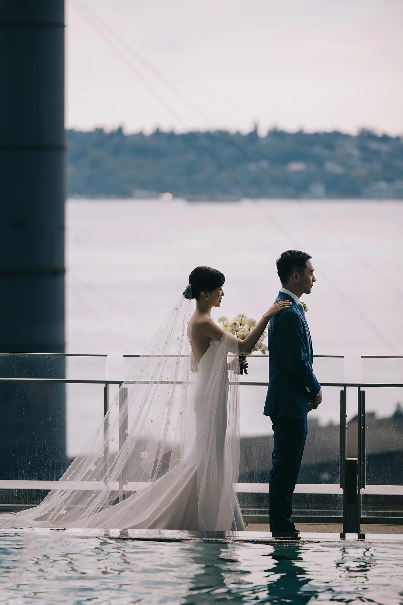 A bride in a white gown and veil places her hand on the shoulder of a groom in a blue suit on a balcony by a body of water, with a distant shoreline in the background.