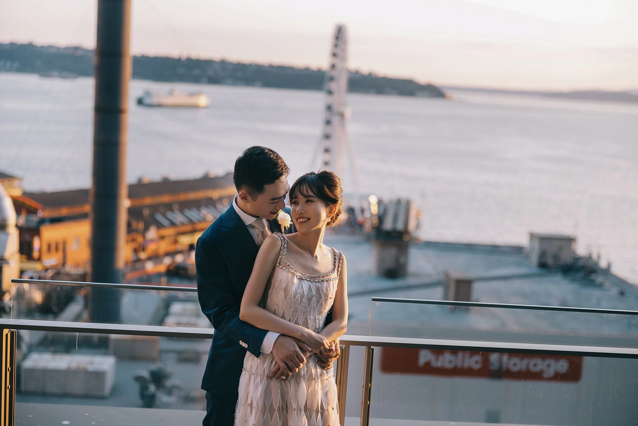 A couple embracing on a balcony overlooking a body of water and a large ferris wheel, with ships and a cityscape in the background during sunset.