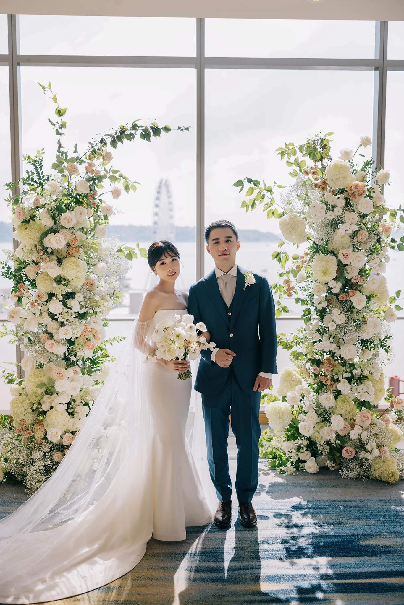 A bride and groom standing in front of floral arrangements and a large window with a city view, including a Ferris wheel in the background.