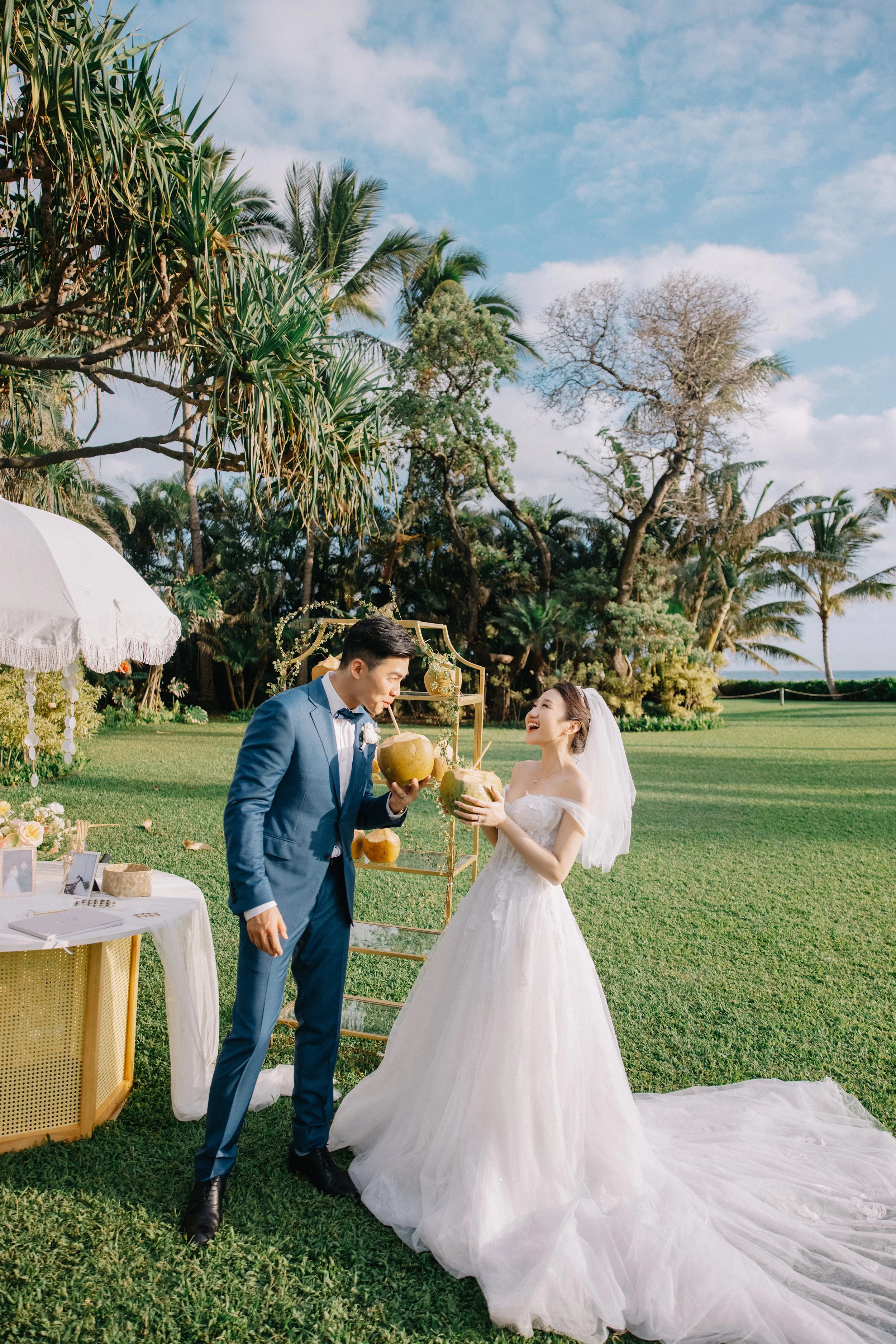 A bride and groom in wedding attire sharing coconuts at an outdoor wedding reception, surrounded by lush greenery and trees under a blue sky.