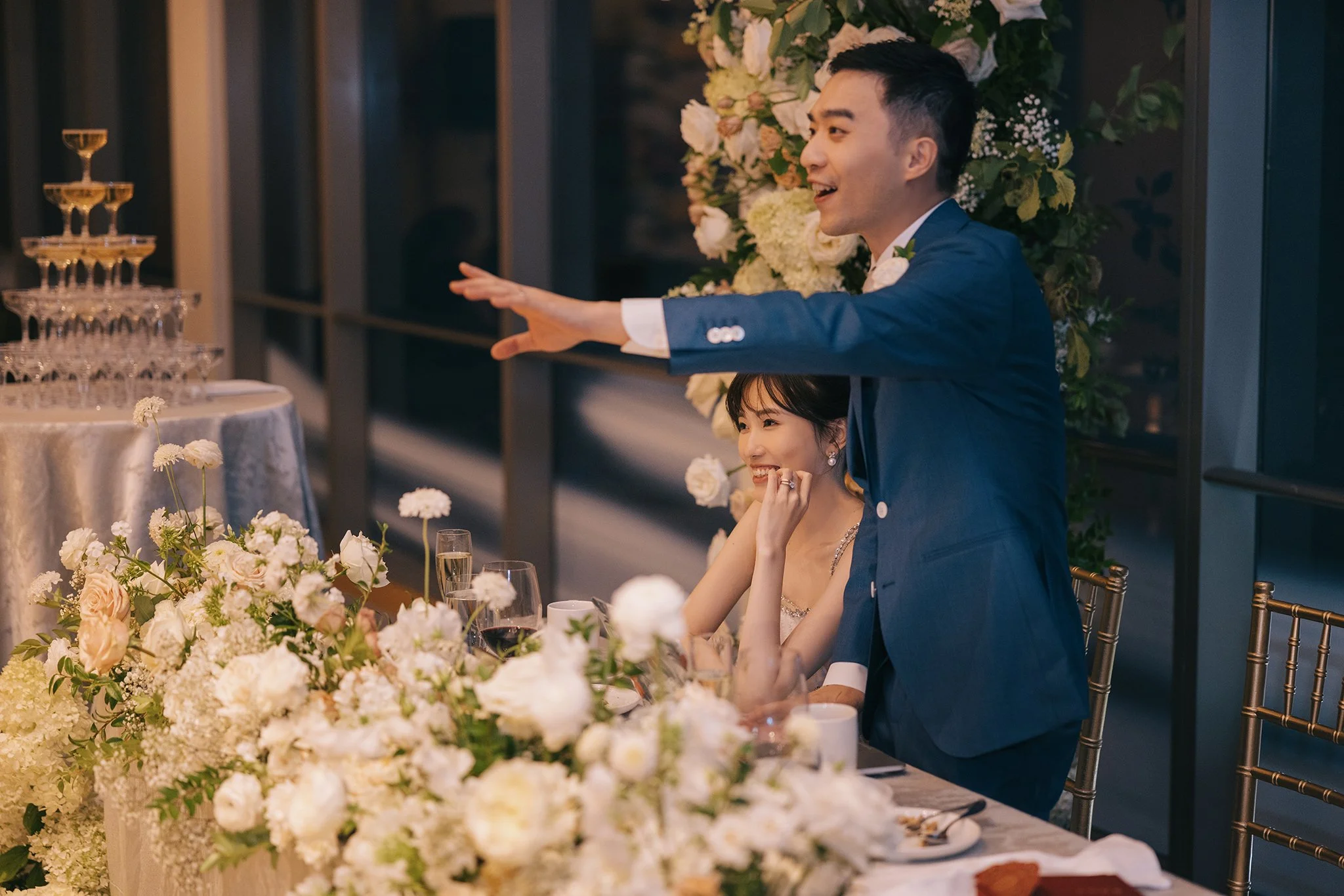 A wedding reception with a groom in a blue suit and a bride in a sleeveless dress, sitting at a table decorated with white and pink flowers. The groom is standing and gesturing with his right arm extended, while the bride is smiling and resting her c