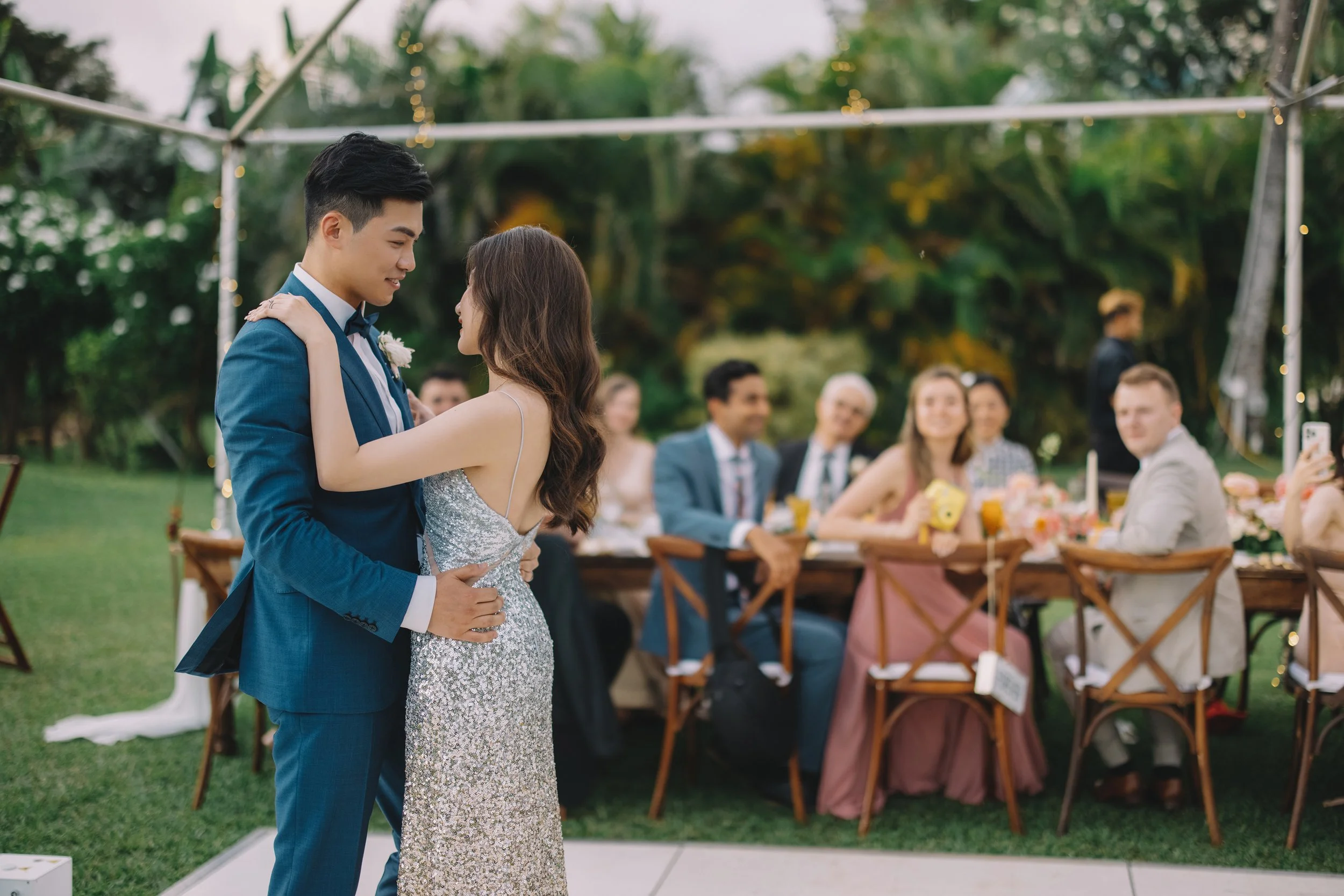 A couple dancing at a wedding reception outdoors, with guests seated at a decorated table in the background.