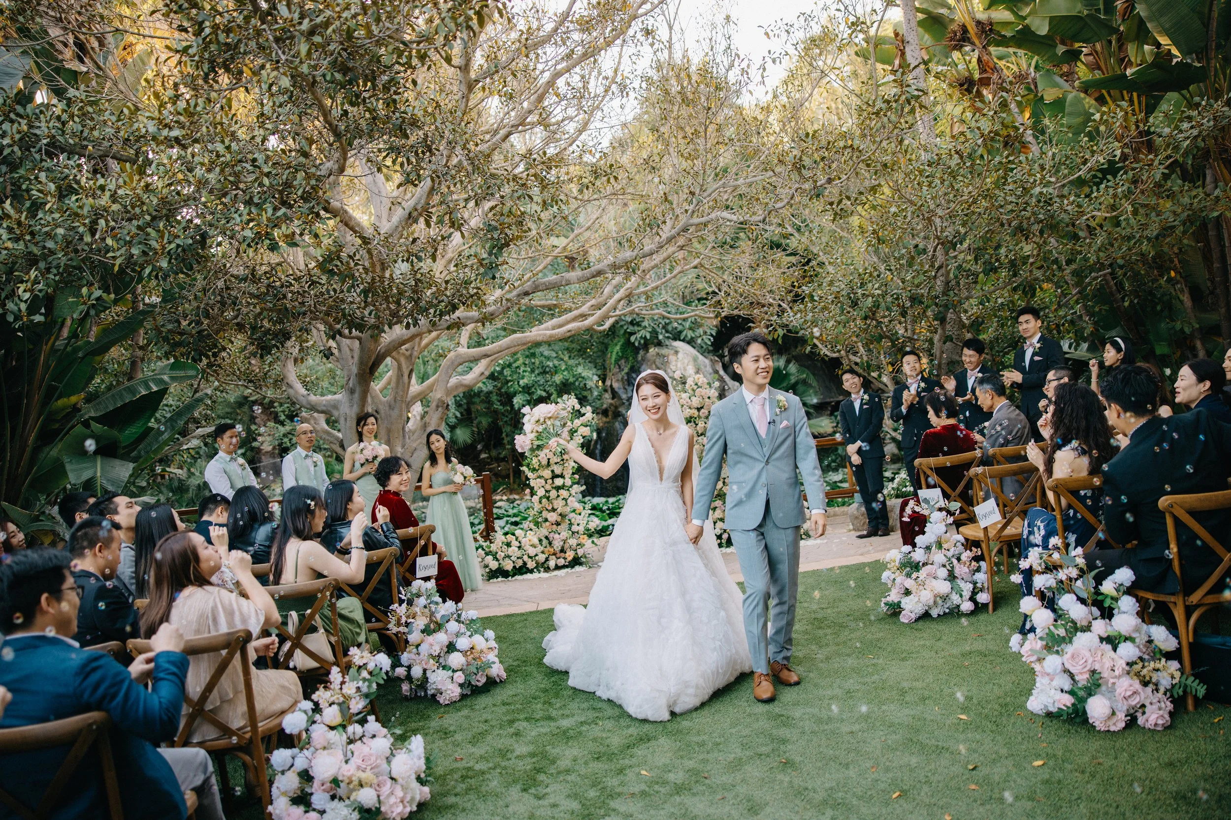 A bride and groom walking hand-in-hand down an outdoor aisle at their wedding, surrounded by guests seated on sides, with trees and floral decorations in the background.