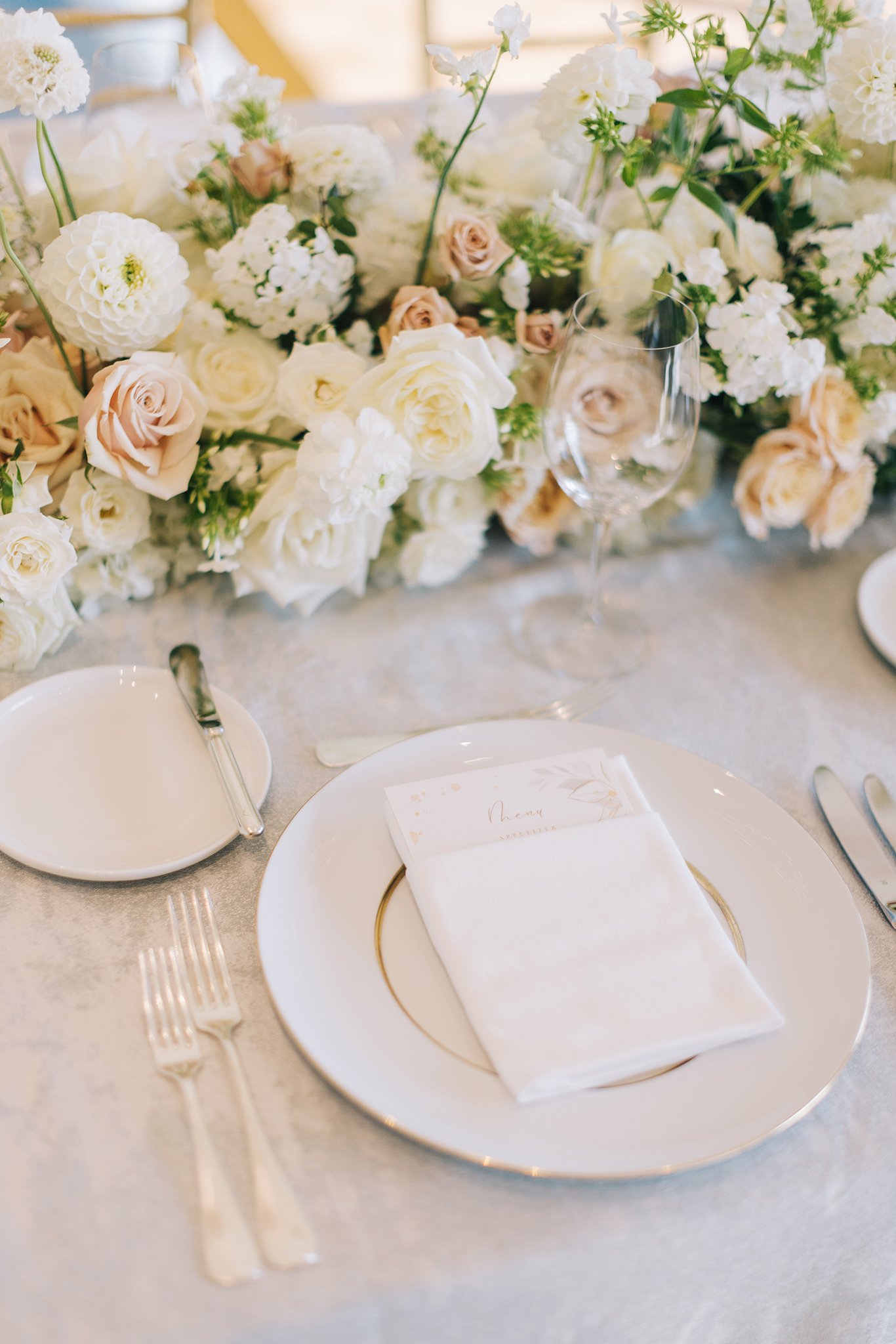 Elegant wedding table setting with a floral centerpiece of white and blush roses, dahlias, and greenery, a white plate with a gold-trimmed charger, a folded napkin, a menu card, and glassware.