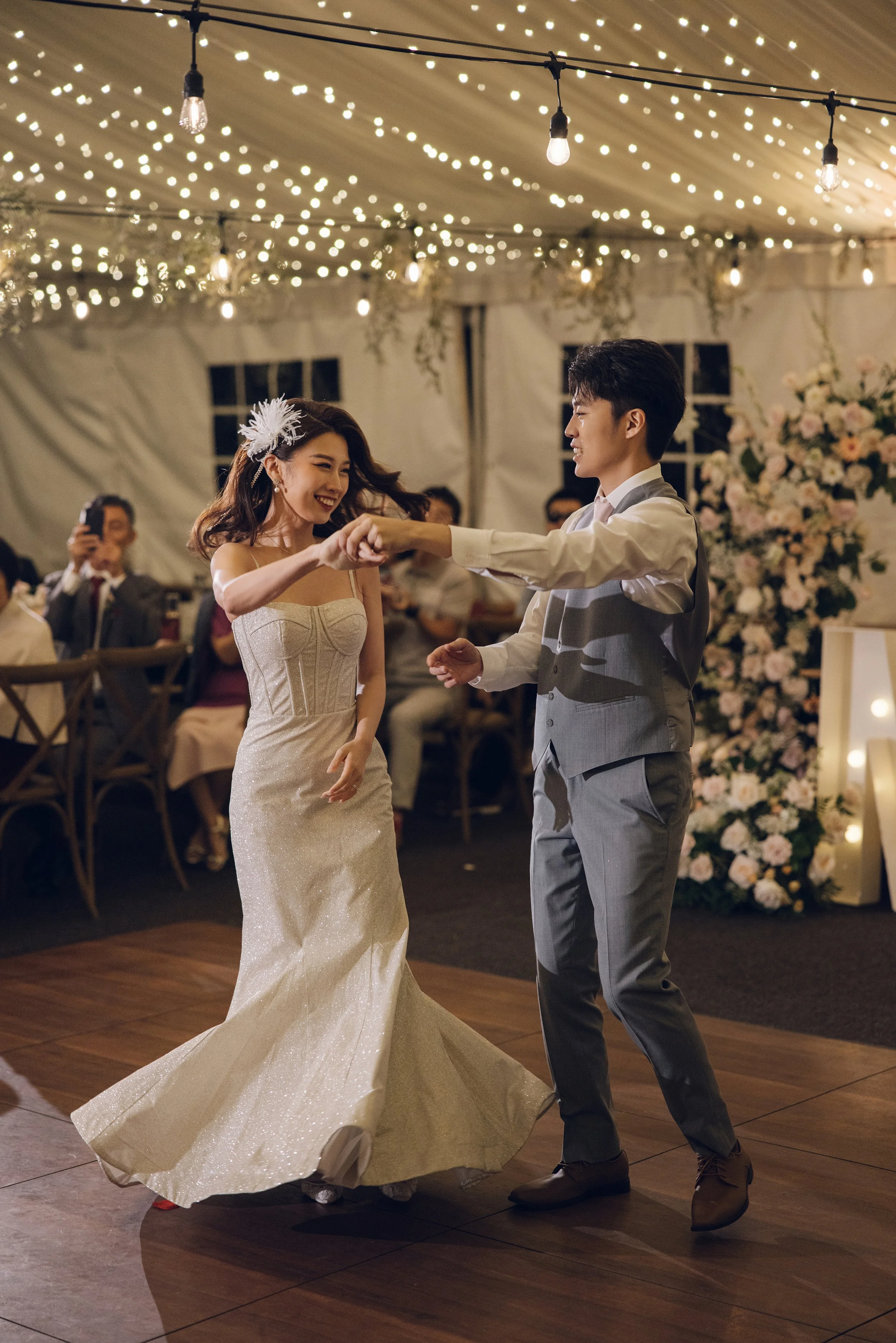 A bride and groom dancing at their wedding reception under string lights with guests watching and smiling in the background.