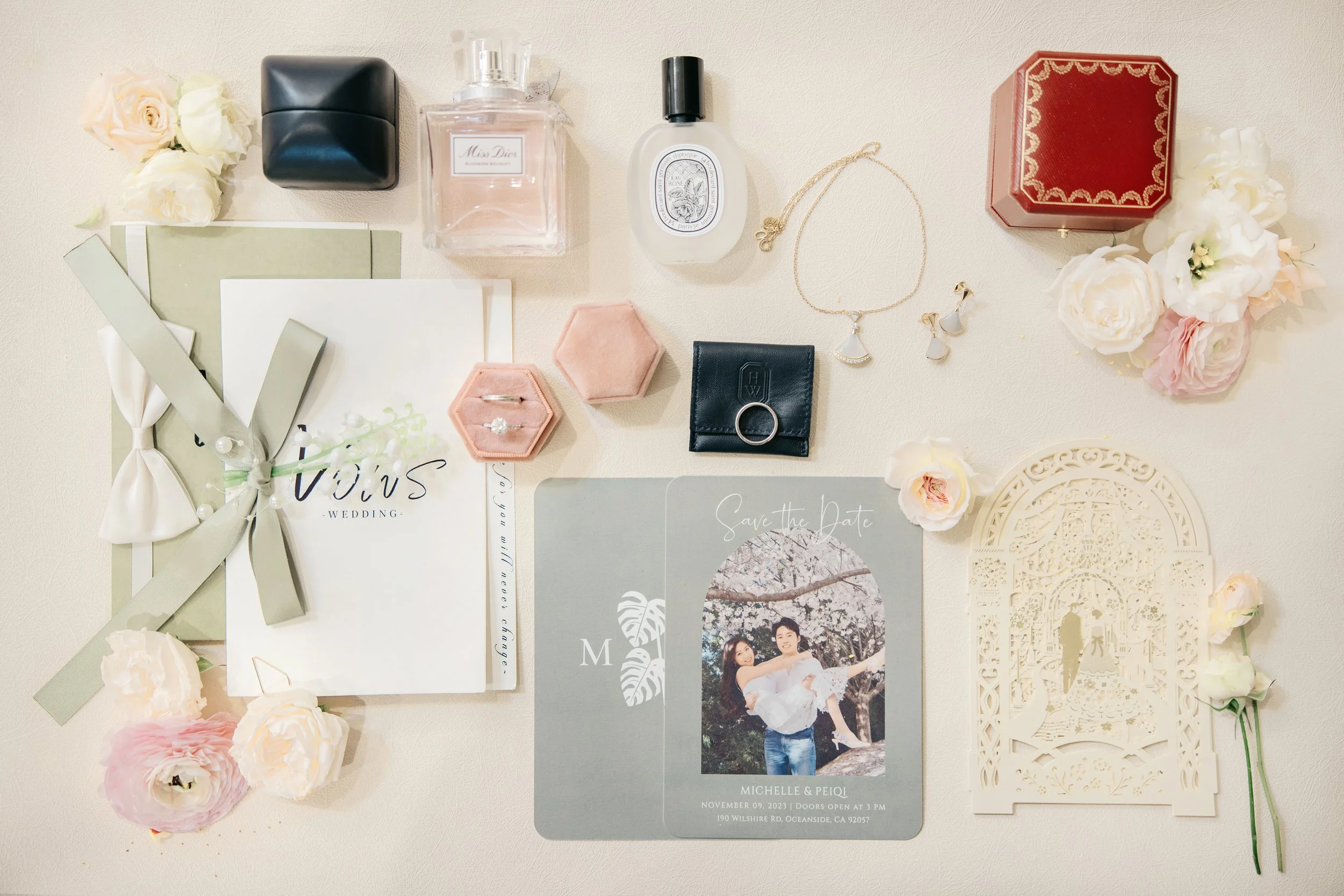 Wedding invitation suite with a photo of a couple, surrounded by flowers, jewelry, perfume, and decorative boxes on a white surface.