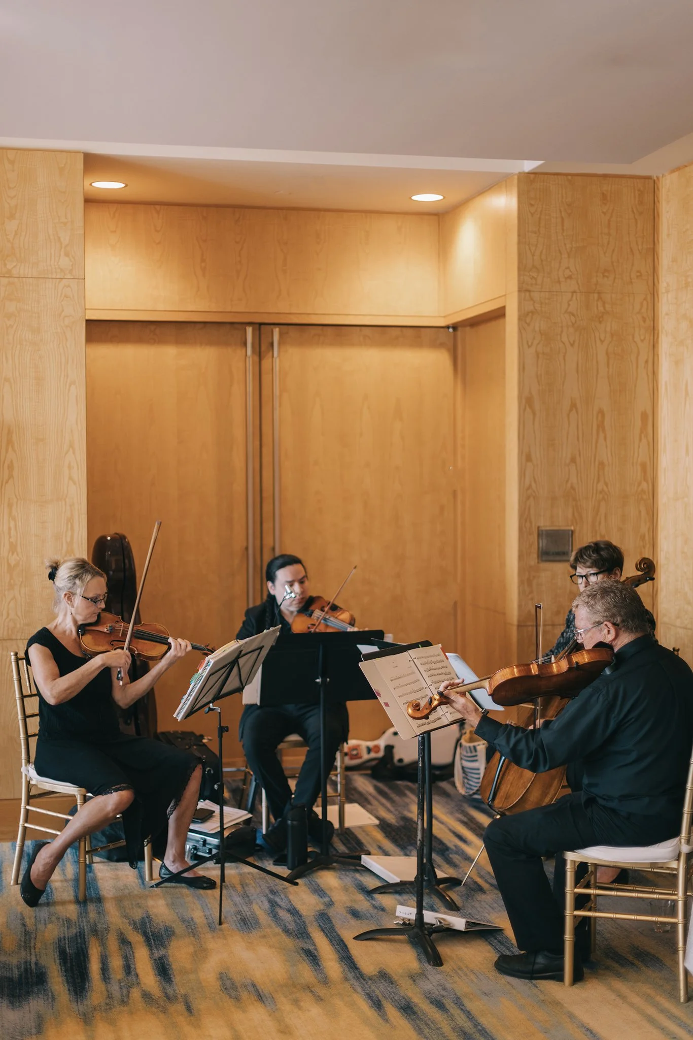 Four musicians playing violins and a cello indoors with wood-paneled walls, music stands, and sheet music.