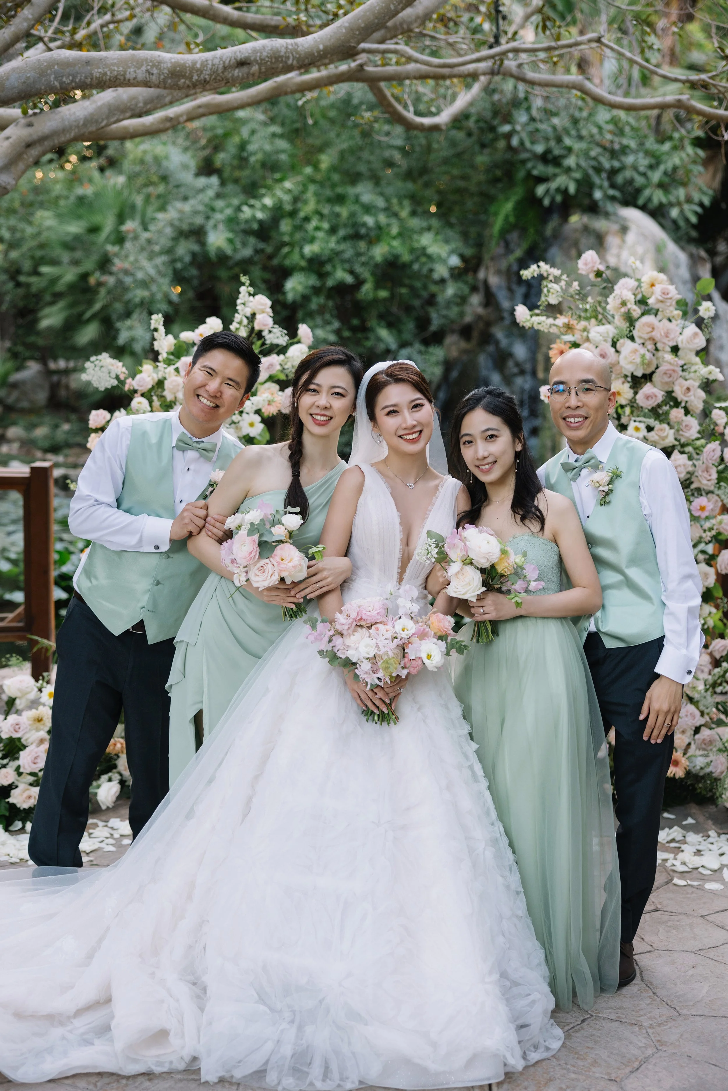 A group of six people, including a bride in a white gown and groom in a suit, standing outdoors among flowers and greenery, smiling at the camera.