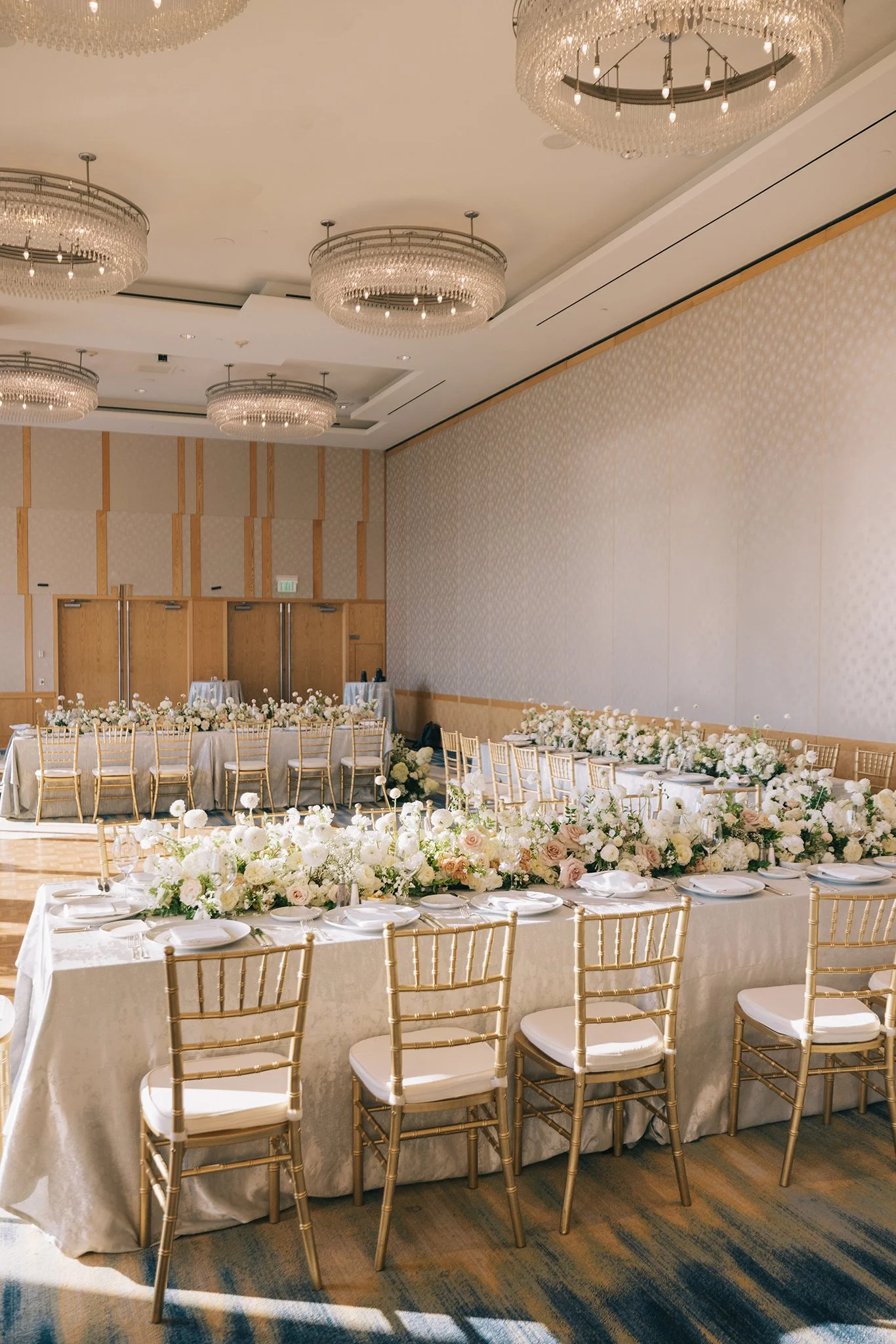 Elegant event hall with long dining tables decorated with white and blush pink flowers and white tableware, surrounded by gold chairs, under crystal chandeliers.