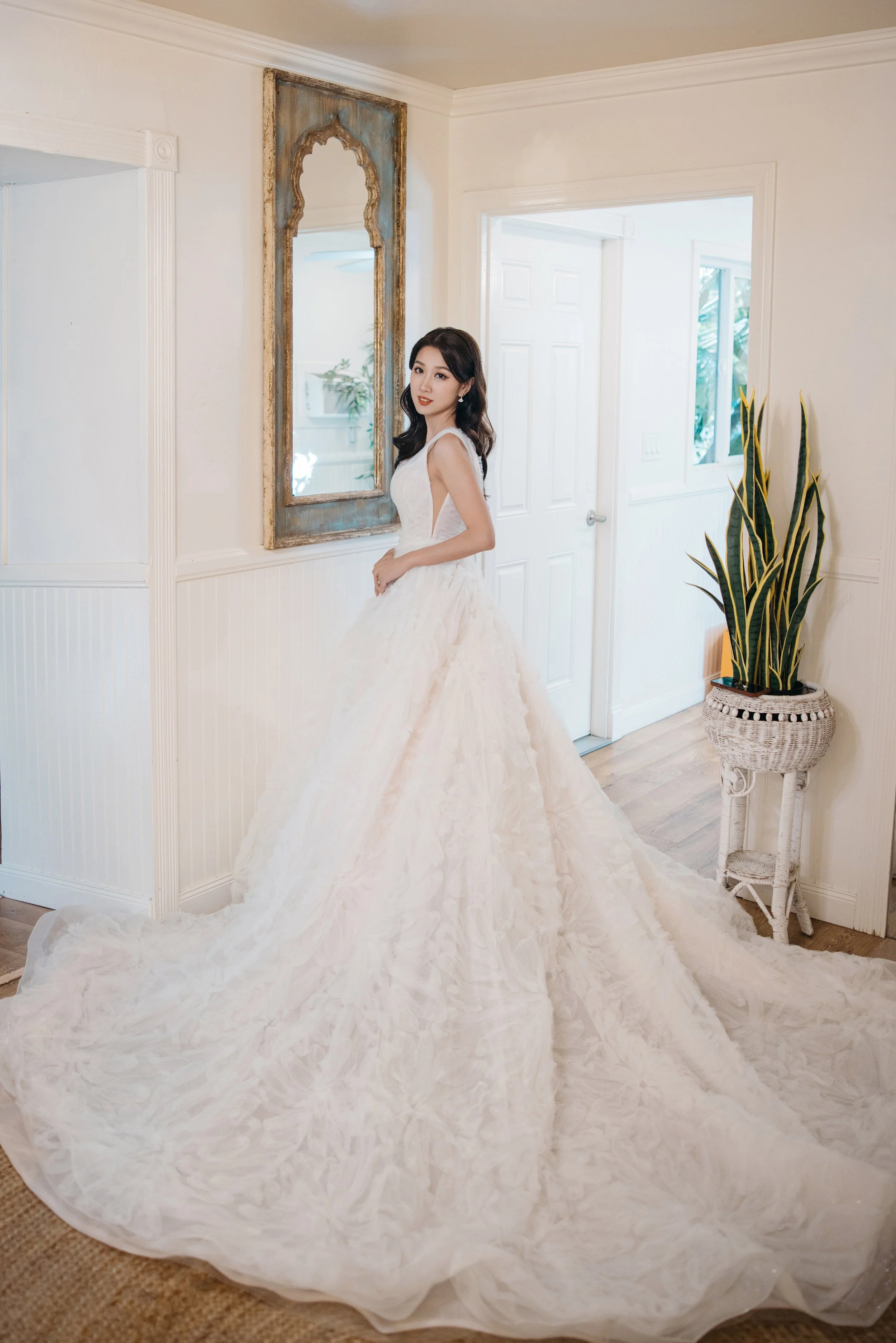 A woman in a white wedding dress standing inside a room with white walls, a mirror, a door, a window, and a potted plant.
