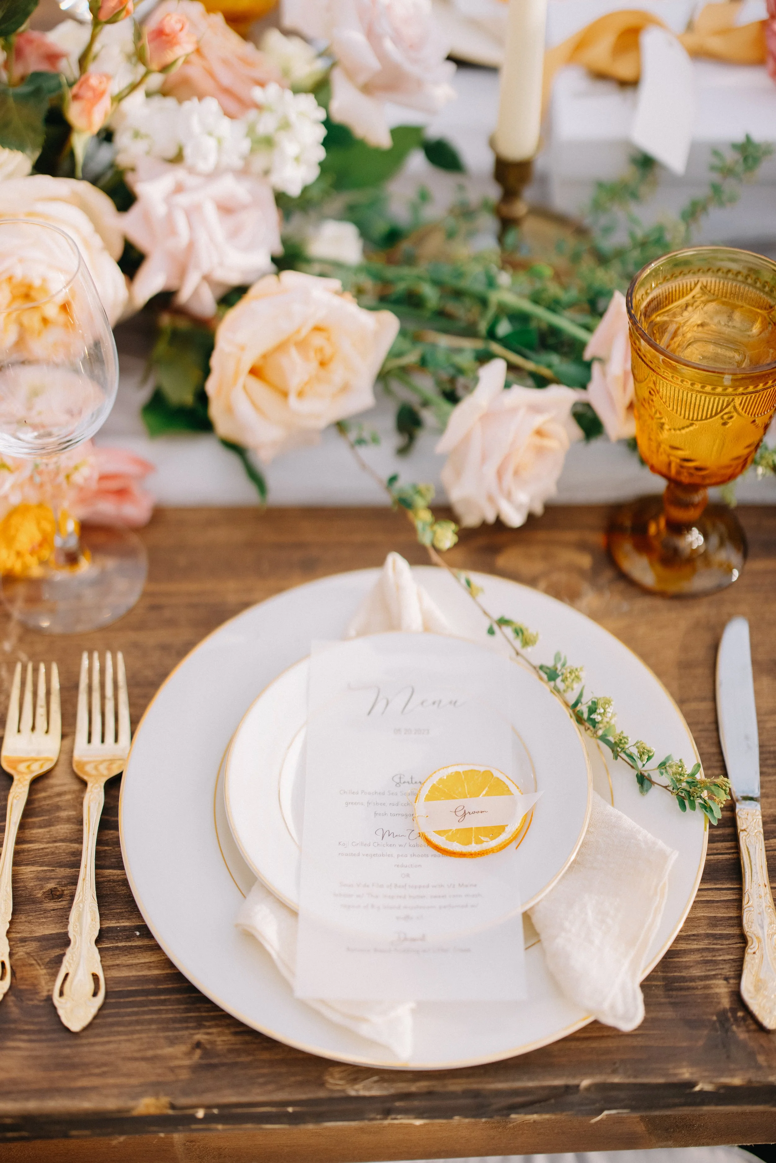 Elegant table setting with a white plate, gold utensils, a menu card with a lemon slice, floral centerpiece with pink and white roses, and amber-colored glasses, on a wooden table with greenery and candles in the background.