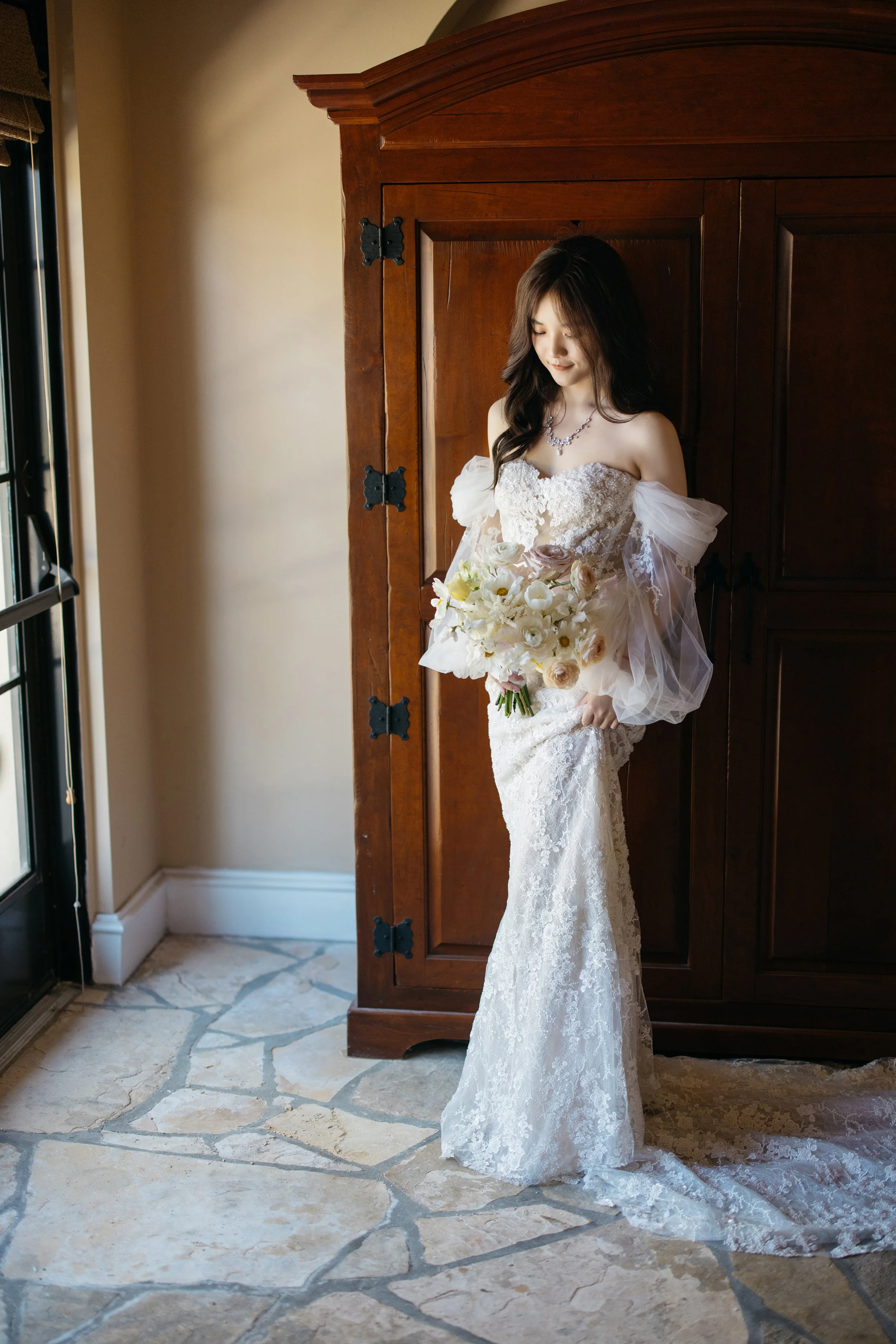 A bride standing indoors near a wooden wardrobe, holding a bouquet of flowers, wearing a lace wedding gown with off-the-shoulder tulle sleeves, looking down and smiling softly.