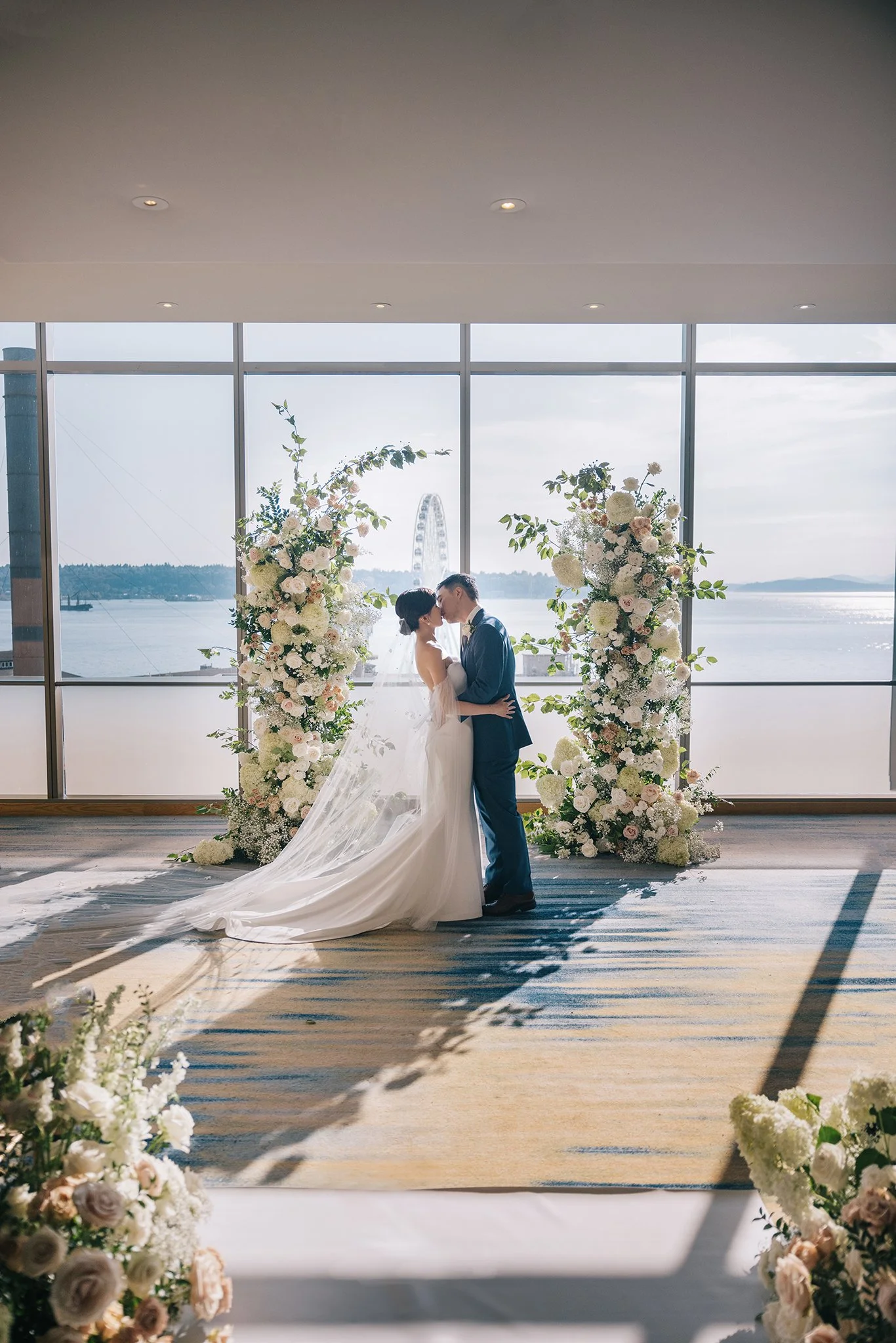 A bride and groom sharing a kiss during their wedding ceremony in front of a large window overlooking a body of water and a Ferris wheel, with floral arrangements on either side.