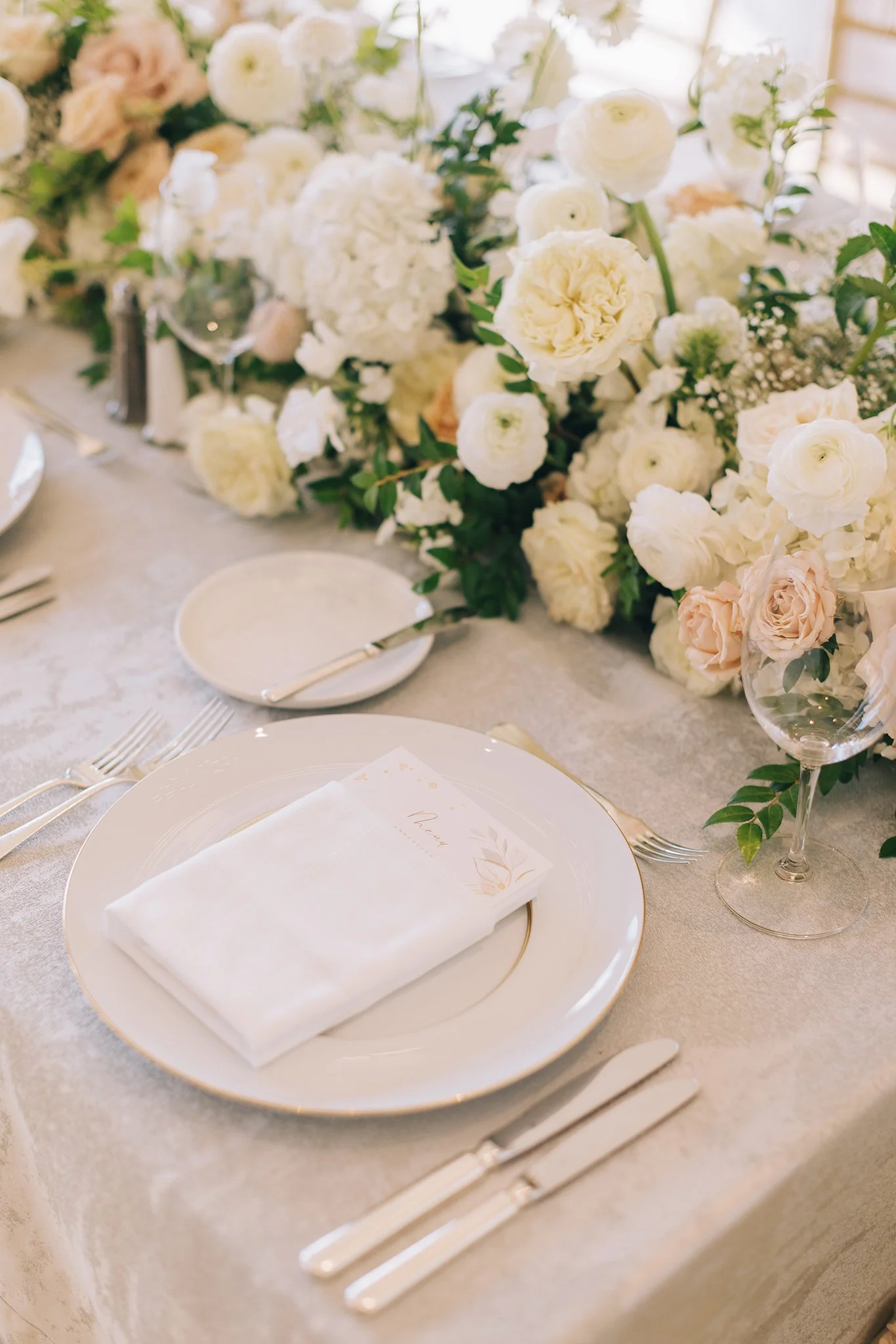 Elegant table setting with a white plate, gold-edged cutlery, a folded white napkin, and a floral arrangement of white and blush roses, hydrangeas, and greenery as a centerpiece.