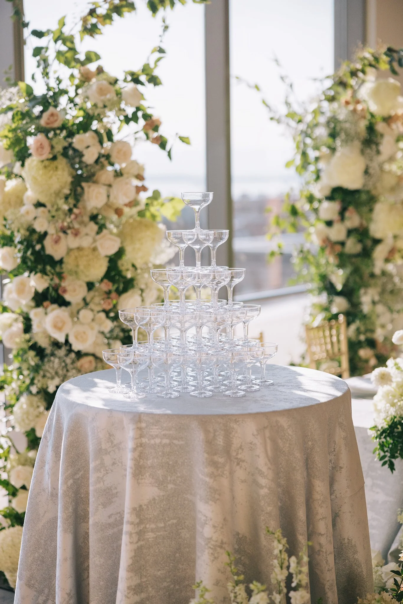 A pyramid of empty champagne glasses arranged on a round table with a white tablecloth, surrounded by floral arrangements of white and light pink roses and greenery, with a large window in the background letting in natural light.