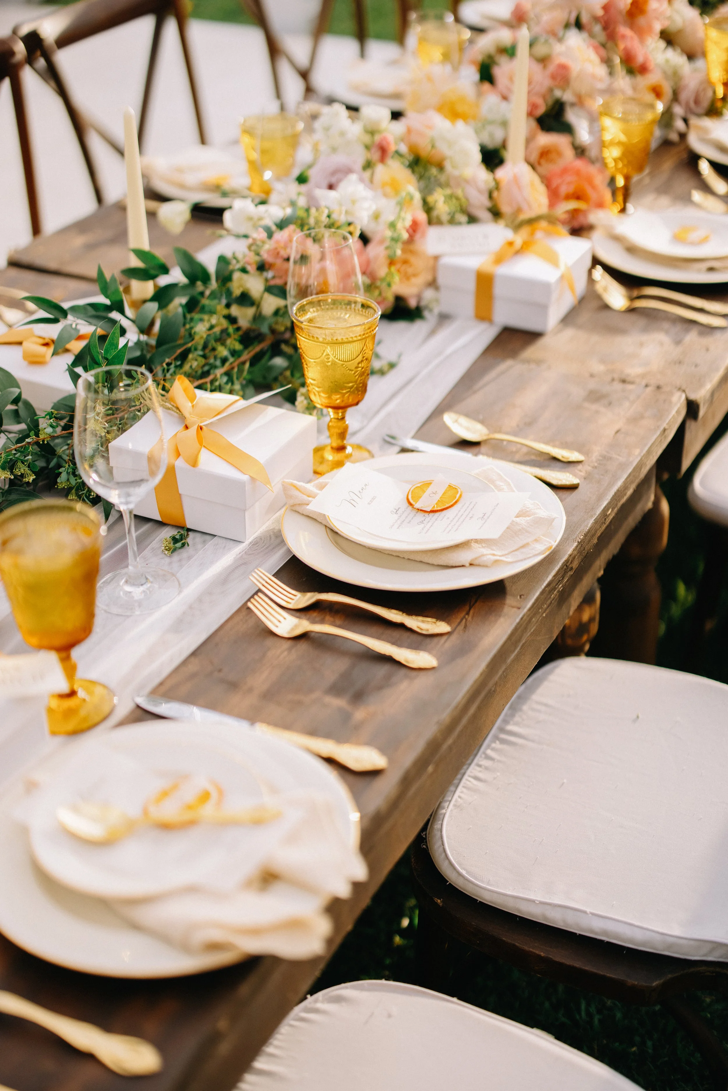 Elegant outdoor dining table decorated with pink and white floral arrangements, with place settings including white plates, gold cutlery, yellow-tinted glassware, and white gift boxes with yellow ribbons available for a wedding or celebration.